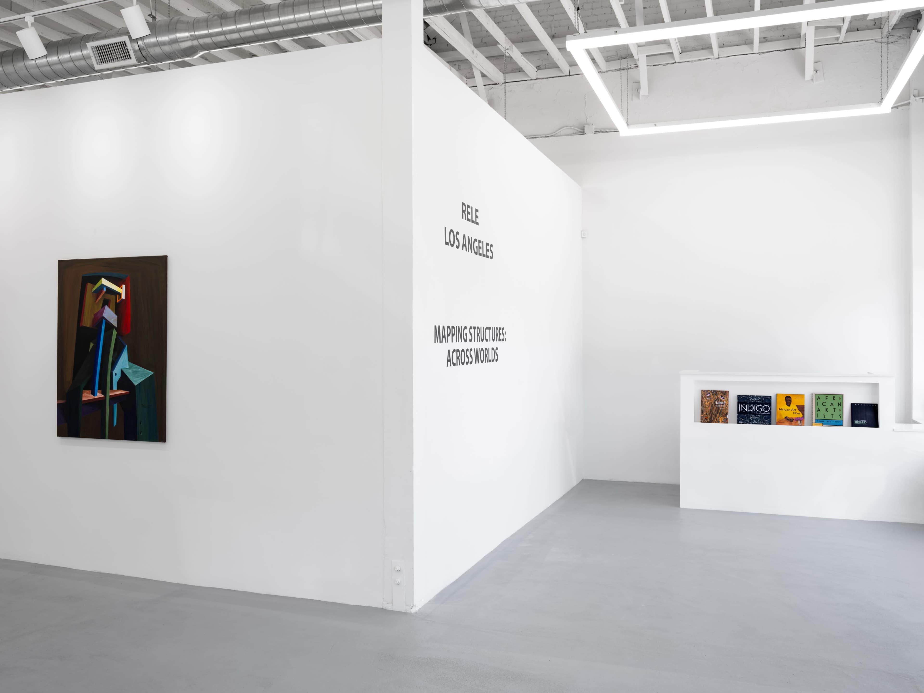 A modern art gallery with a colorful painting on the left wall and a display of books on a shelf against a white wall.