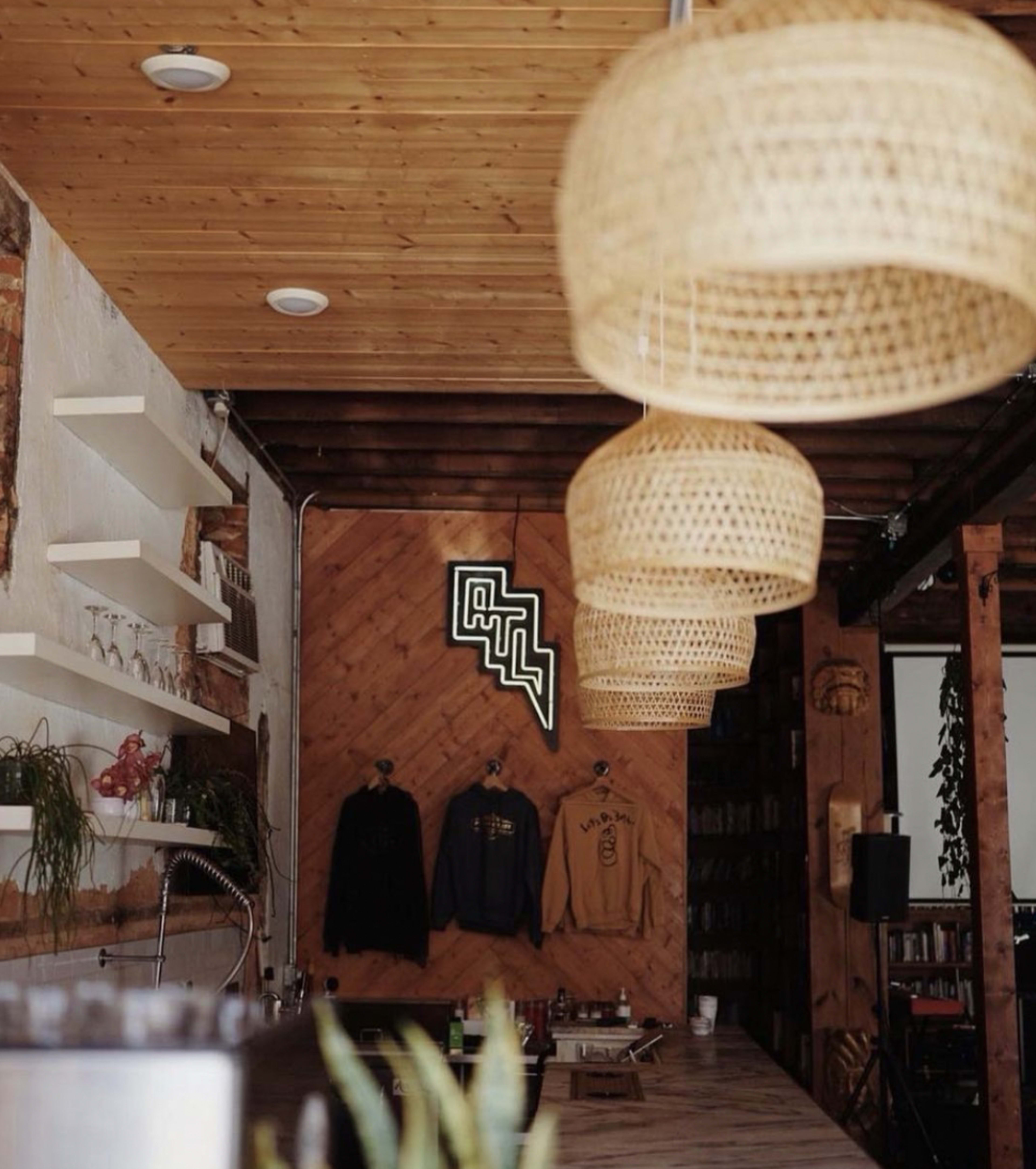 A cozy interior of a café with woven pendant lights hanging from a wooden ceiling, wooden shelves displaying items, and sweatshirts on hangers against a textured wall.