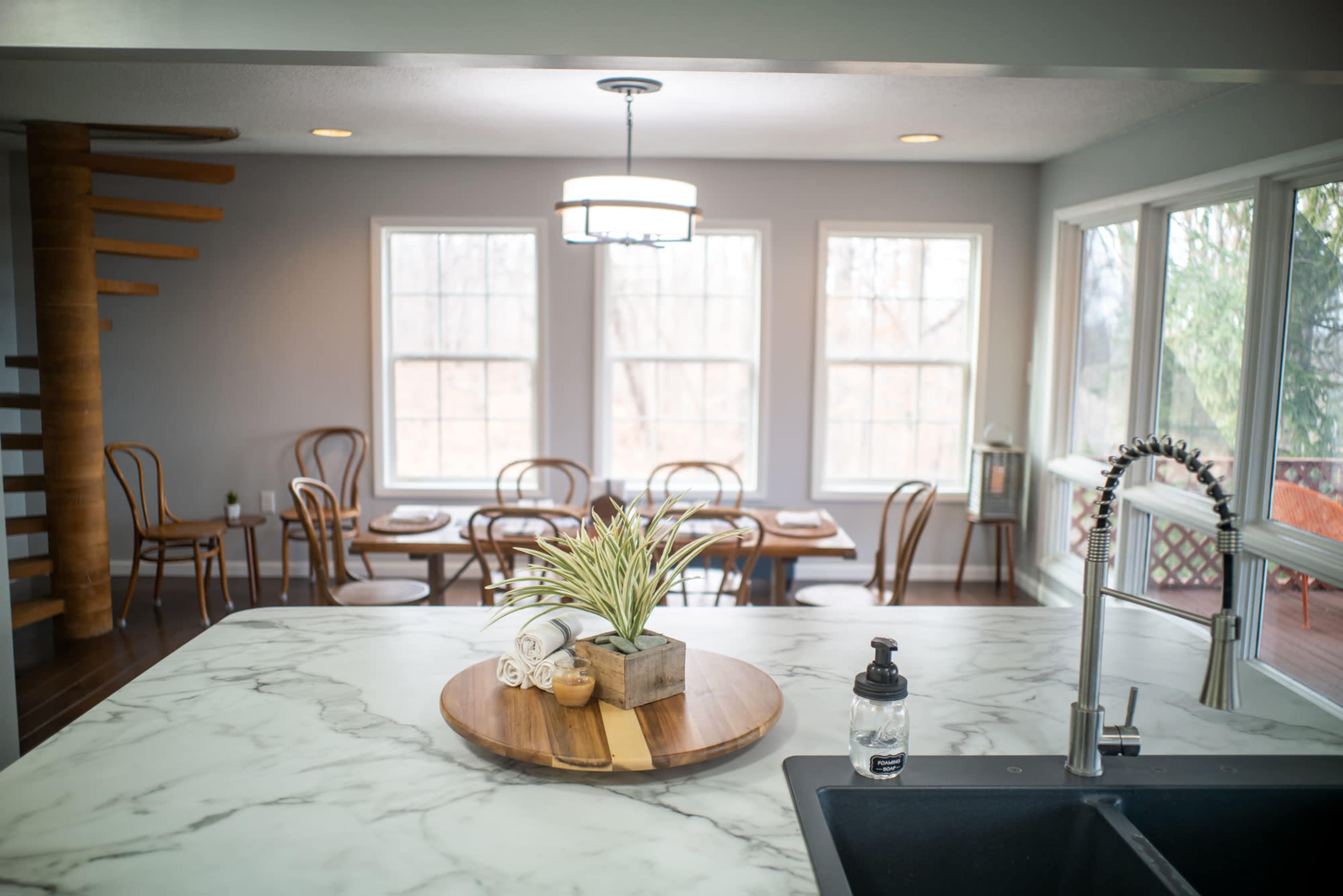 A dining area features several chairs around a table, with large windows providing natural light, and a kitchen island with a marble countertop in the foreground.