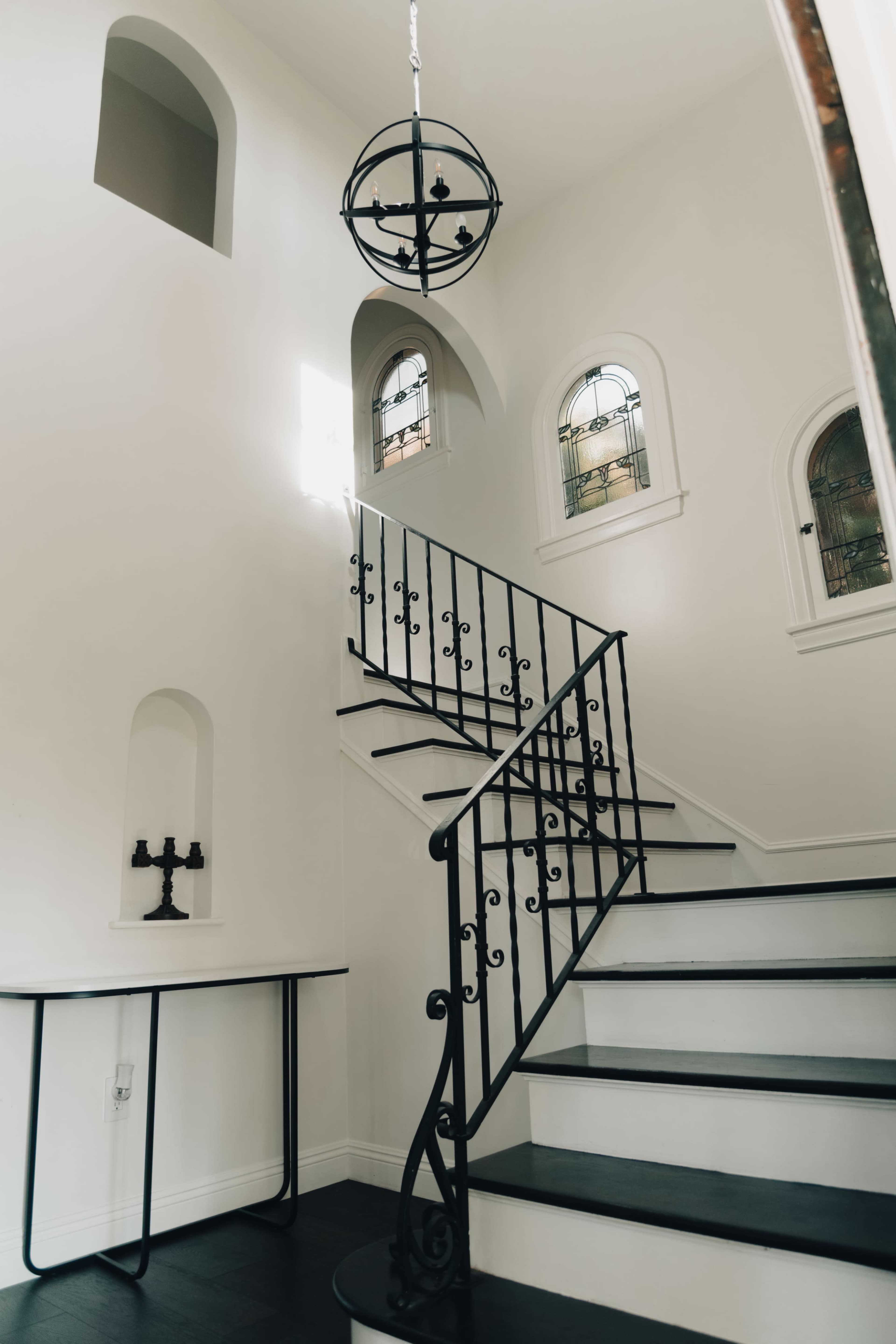 A white staircase with a black wrought-iron railing, a circular chandelier hanging above, and stained glass windows illuminating the space.