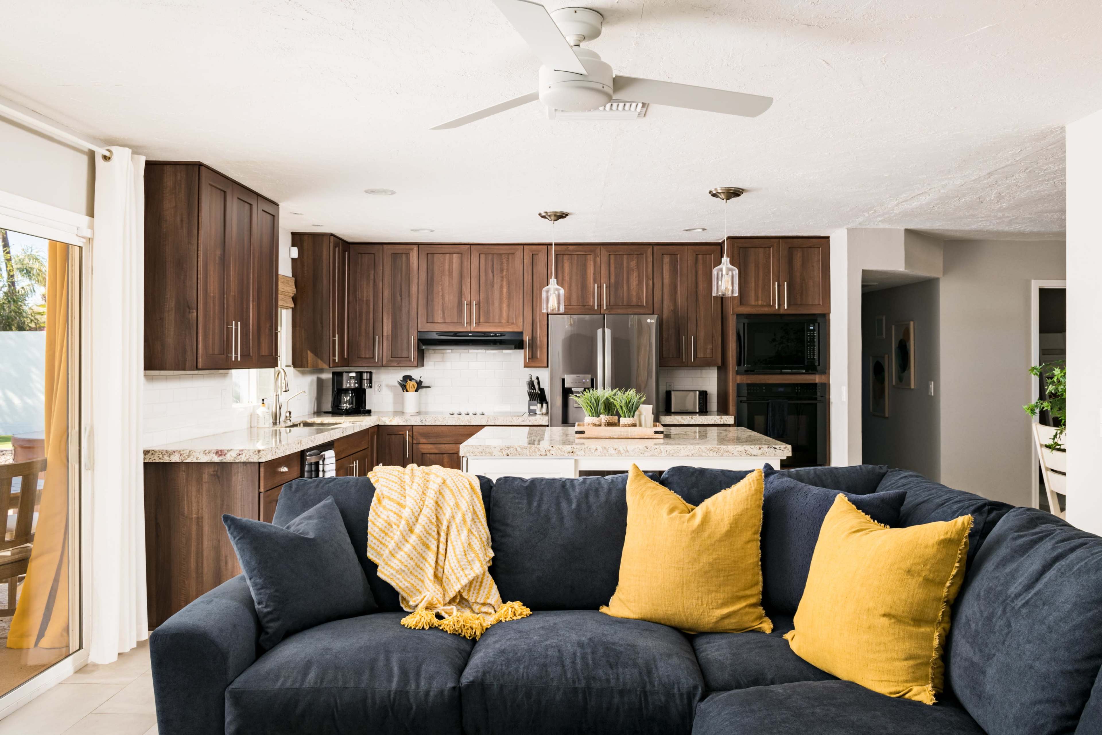 A modern kitchen features wooden cabinets, stainless steel appliances, and a spacious island, with a blue sectional sofa and yellow pillows in the foreground.