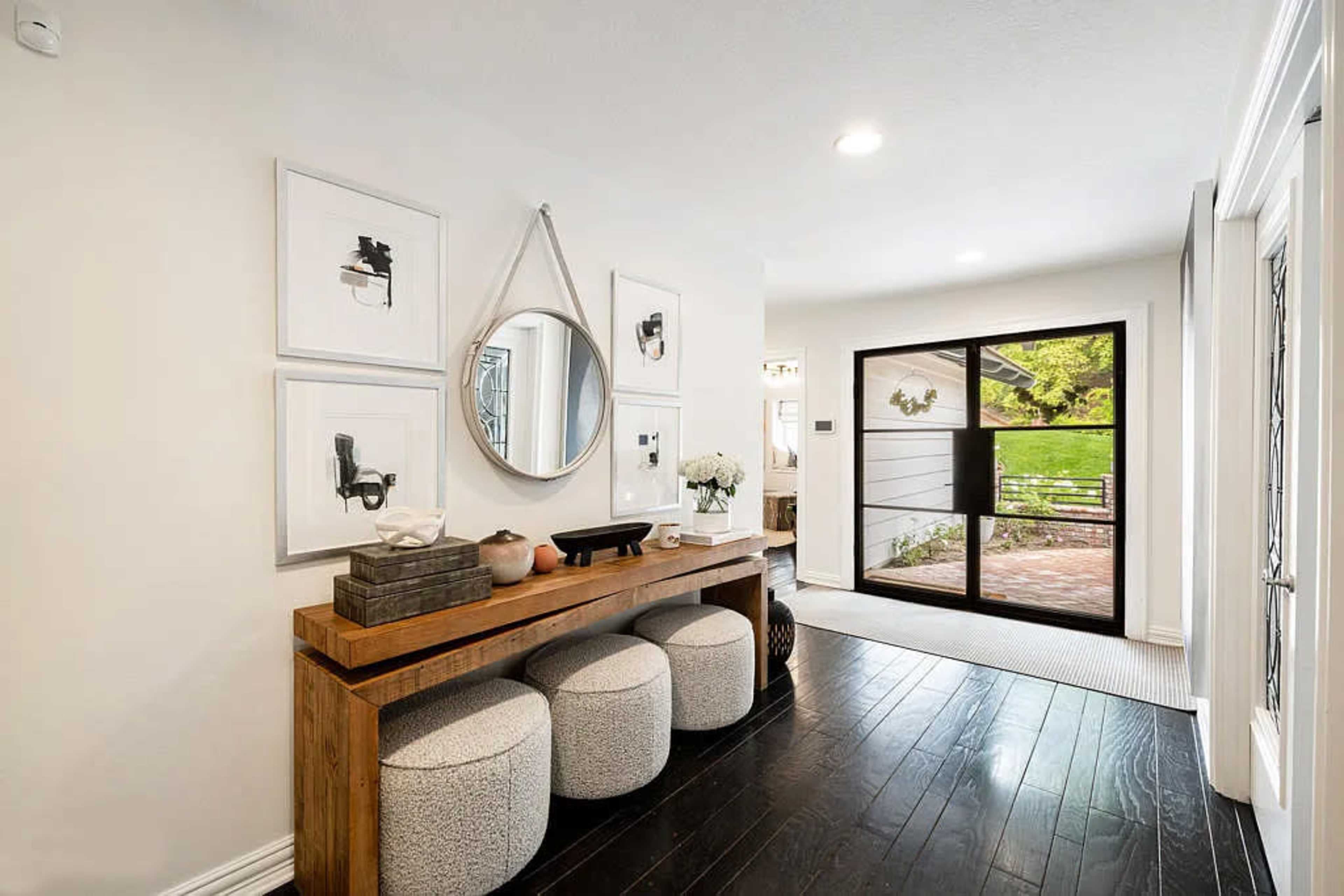 A bright entryway with a wooden console table, decorative stools, and framed artwork, leading to a glass sliding door that opens to a patio.