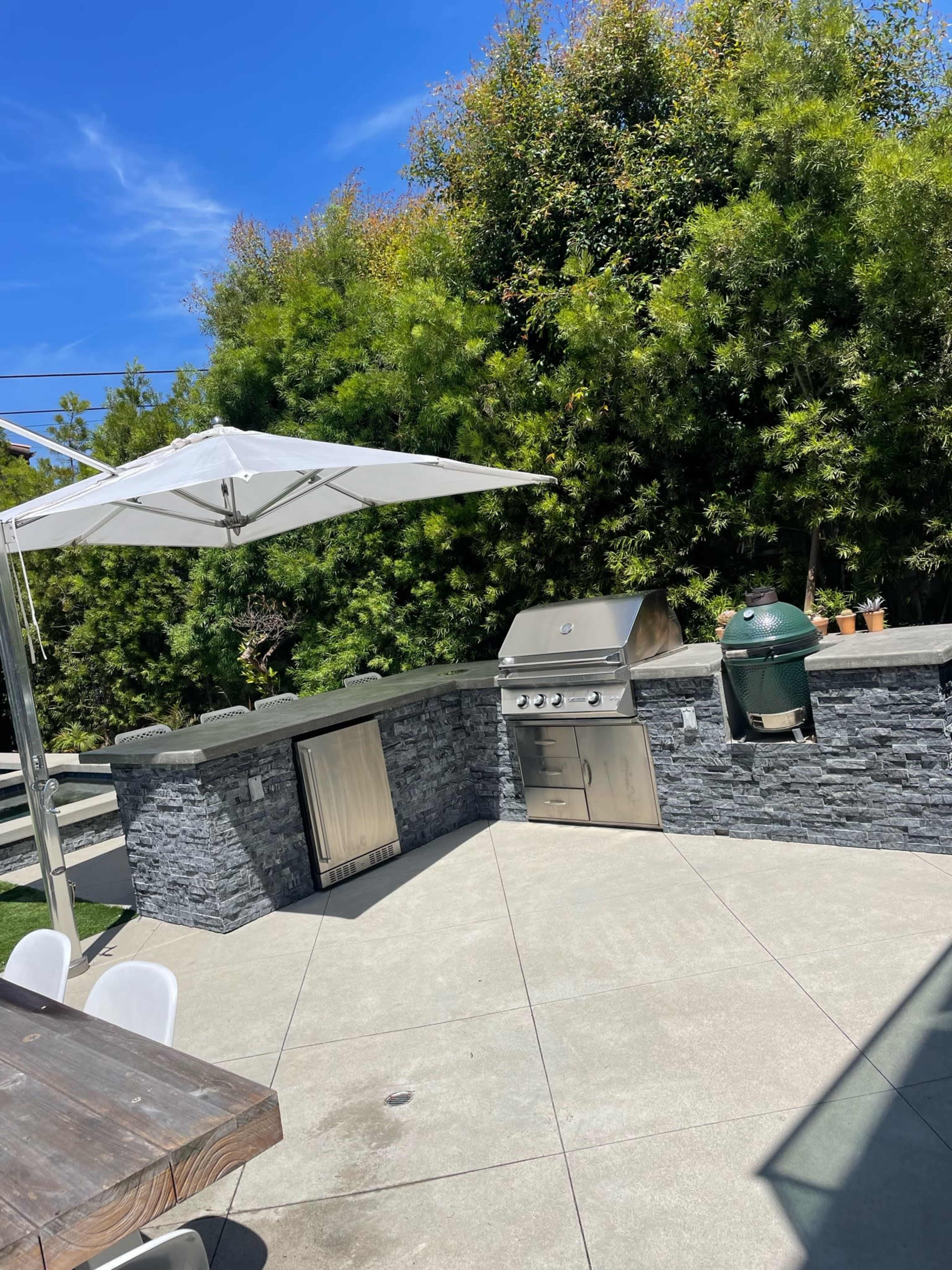 An outdoor patio featuring a stainless steel grill, a stone countertop, an outdoor refrigerator, and a large white umbrella beside a green lawn and bamboo trees.