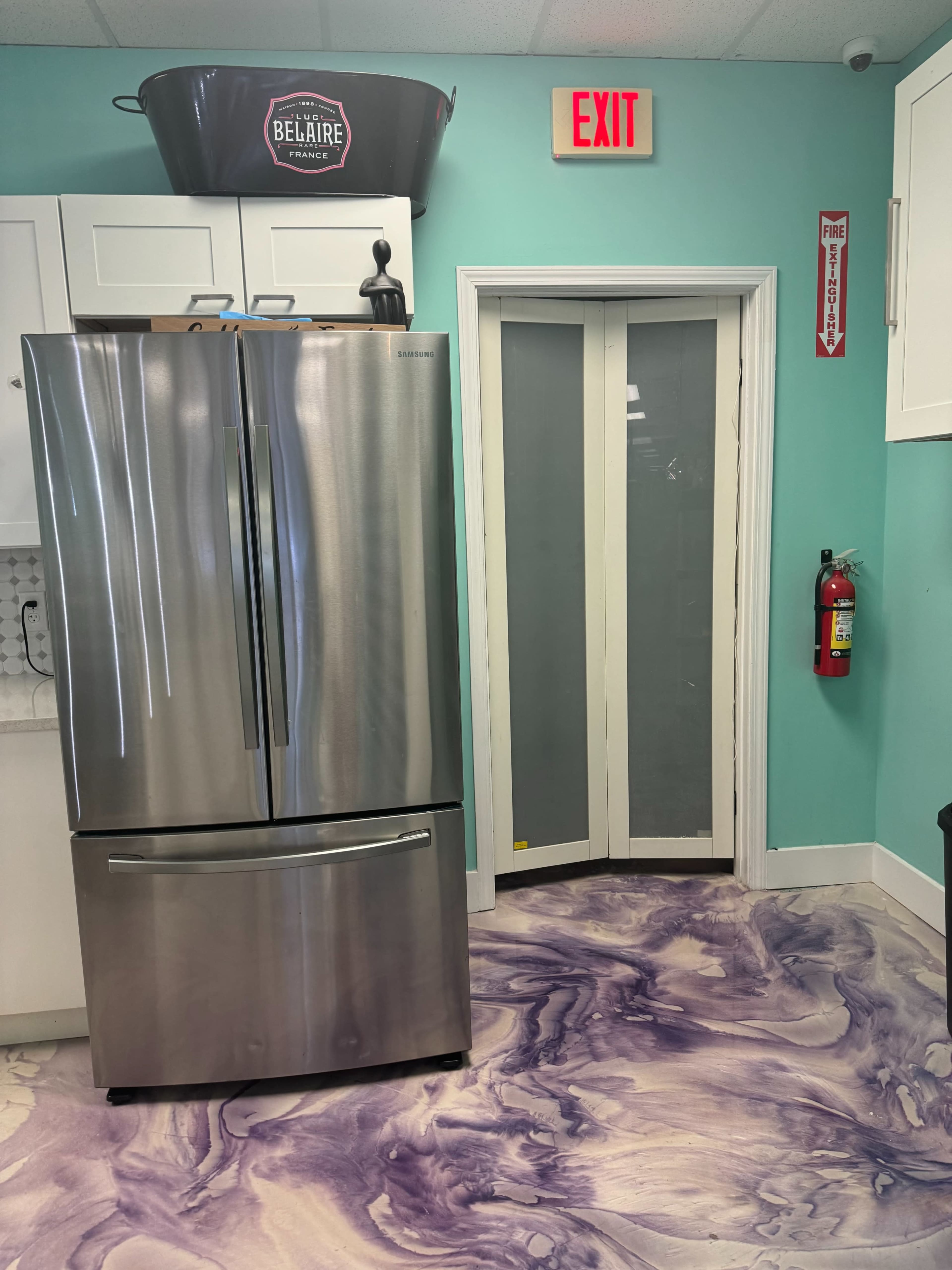 The image shows a kitchen corner with a stainless steel refrigerator, a fire extinguisher on the wall, and a purple marbled floor leading to a door.