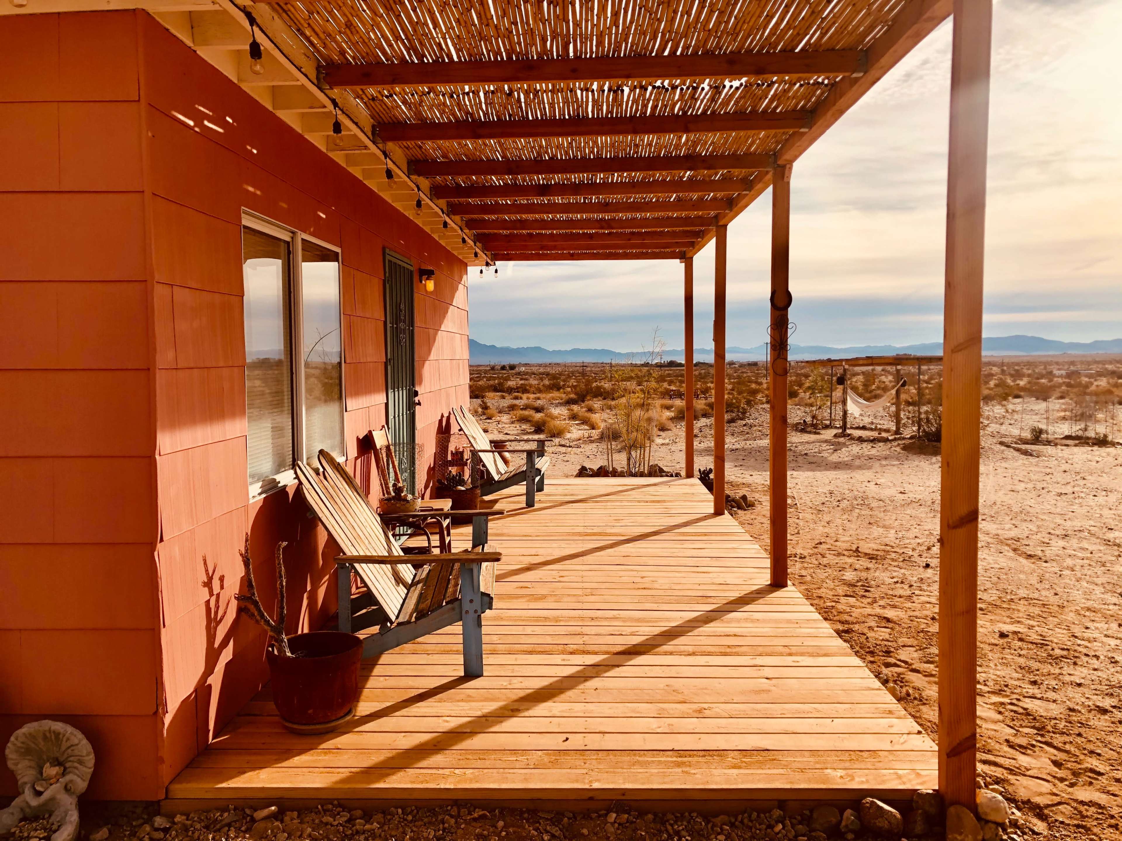 A wooden deck with chairs and potted plants extends from a pink building, overlooking a desert landscape and mountains in the distance.