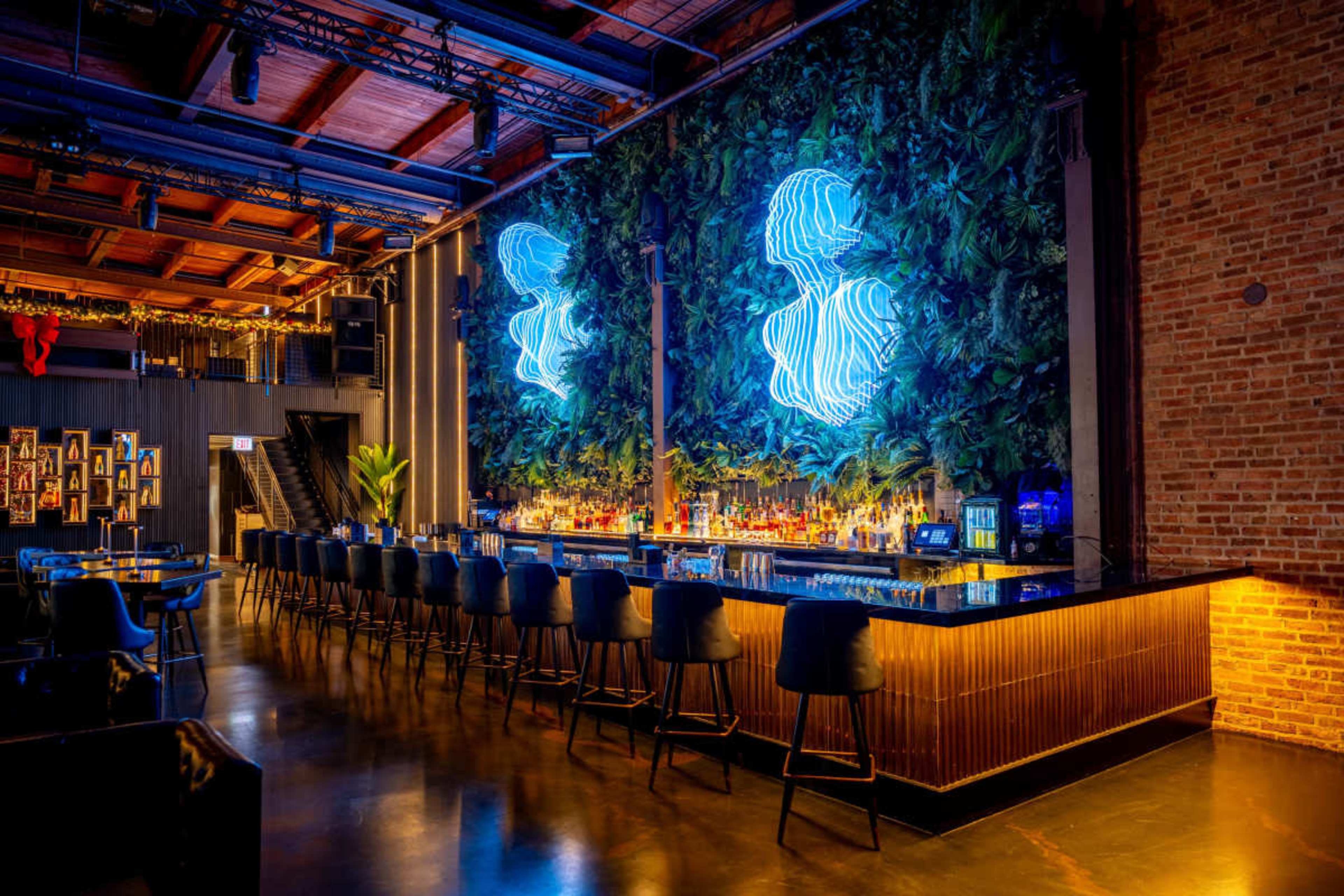 The image shows a modern bar area with a large neon light display against a green wall of plants, featuring sleek bar stools and a polished counter.