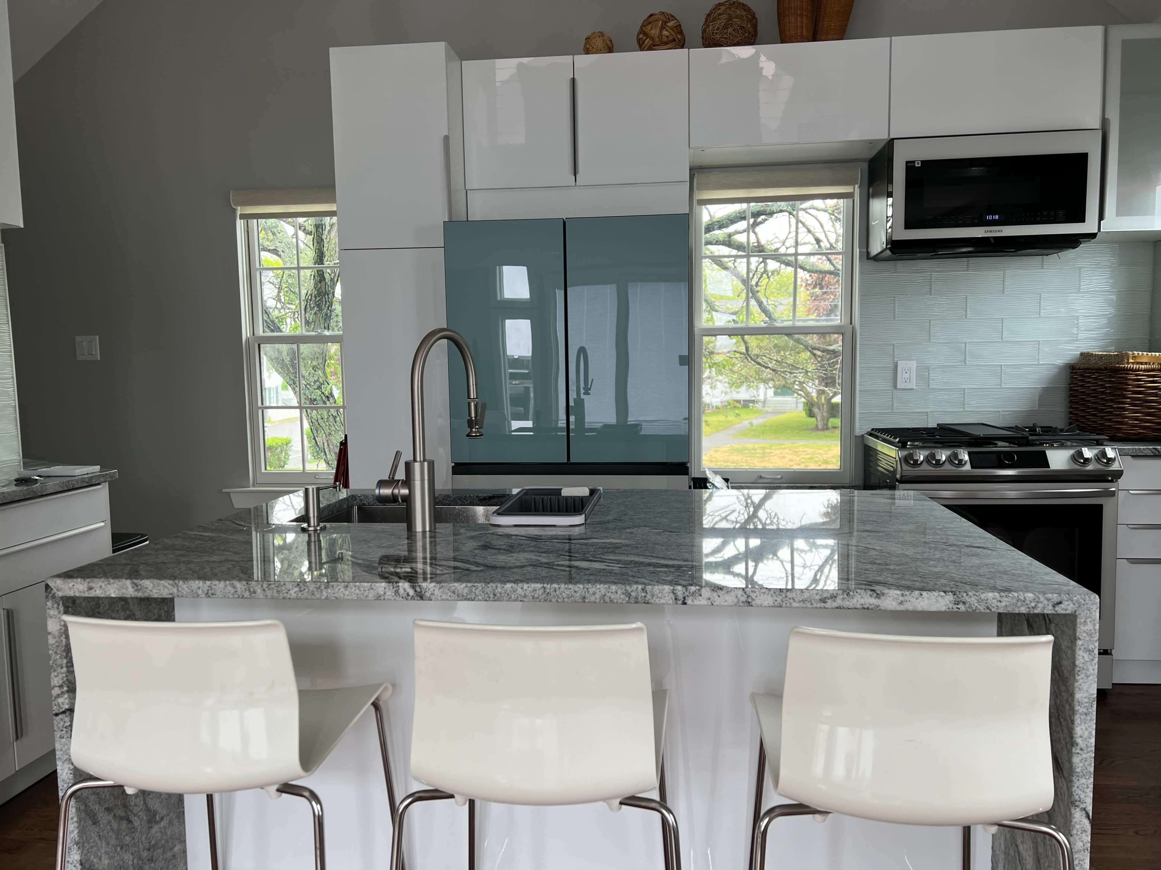 A modern kitchen features a large granite island with three white stools, a blue refrigerator, and large windows overlooking a garden.