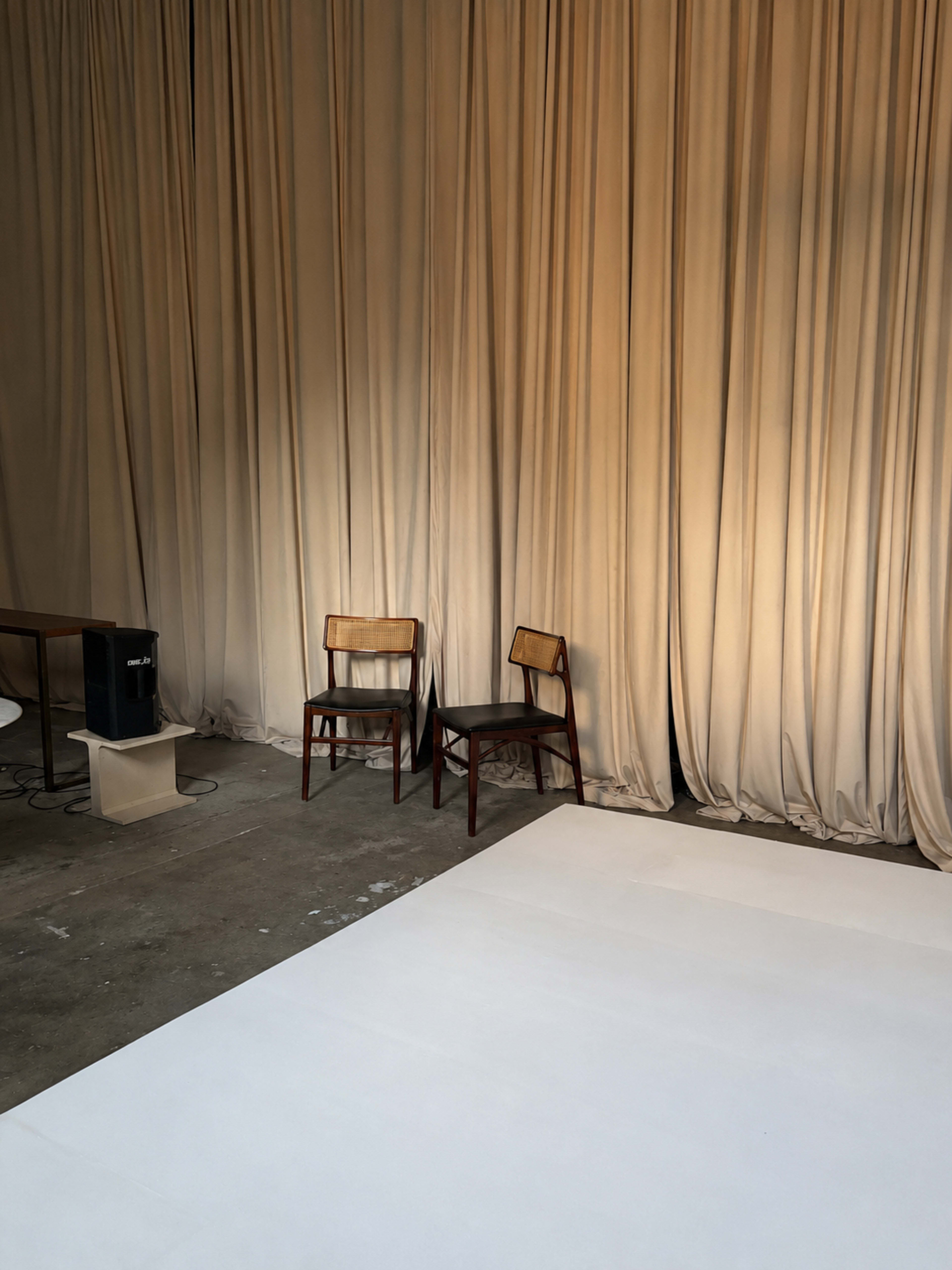The image shows a minimalistic indoor space featuring two wooden chairs near a beige backdrop, with a small table and a speaker positioned nearby on a concrete floor.