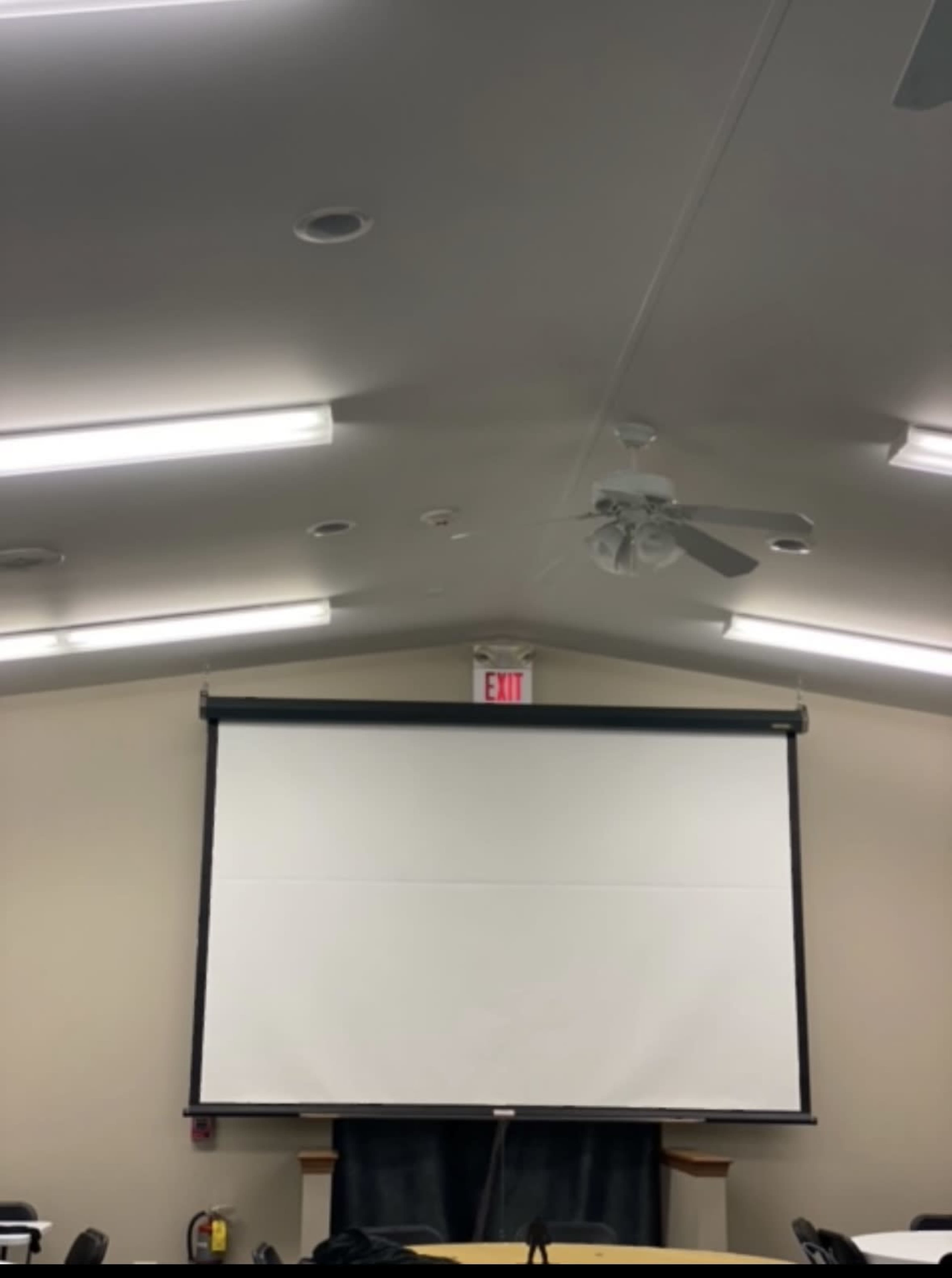 An empty room with a ceiling fan, overhead lights, and a large retractable projector screen mounted on the front wall.