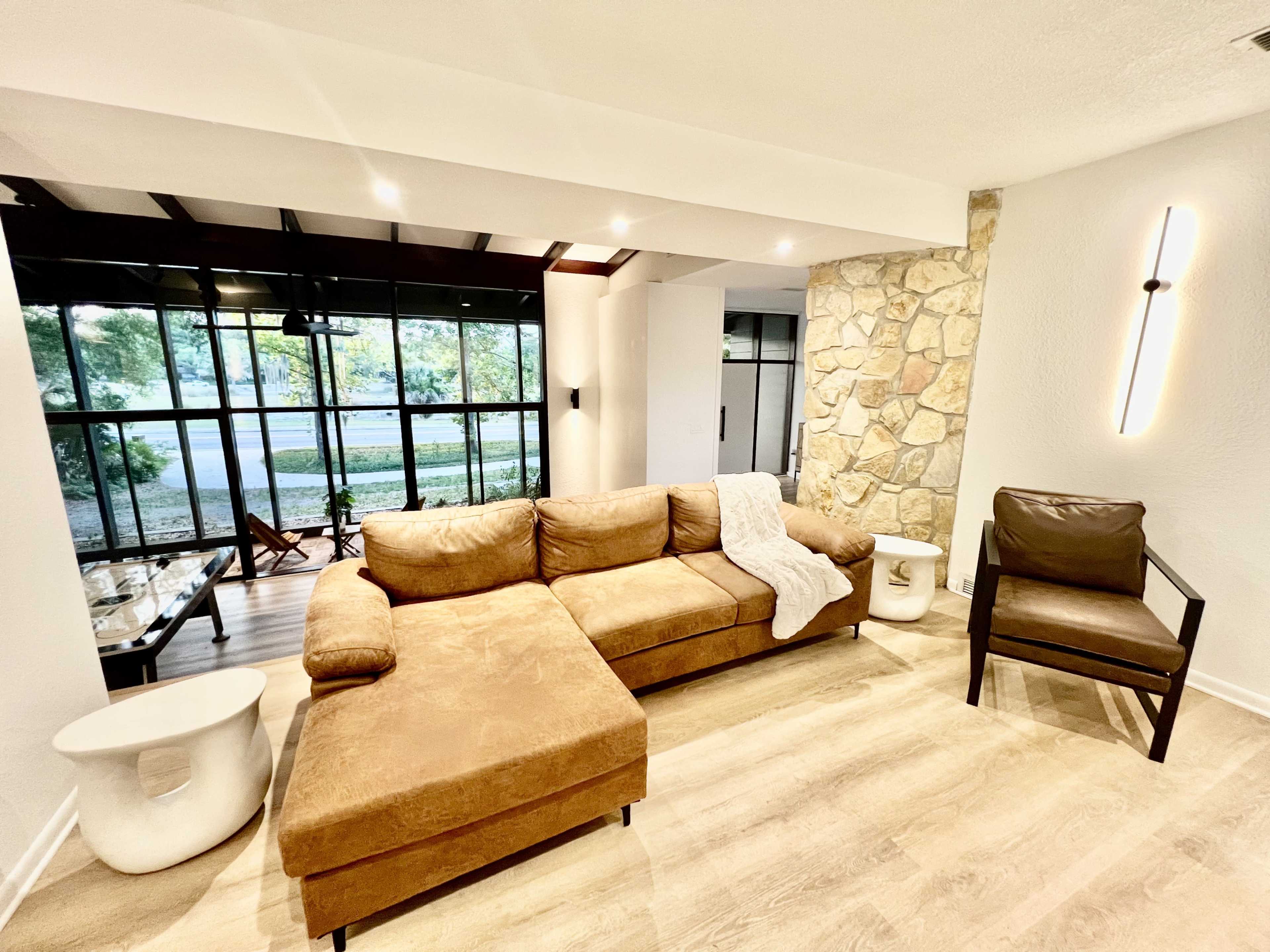 The image shows a modern living room with a brown sectional sofa, a glass coffee table, and a stone wall, along with a leather chair and large windows providing natural light.