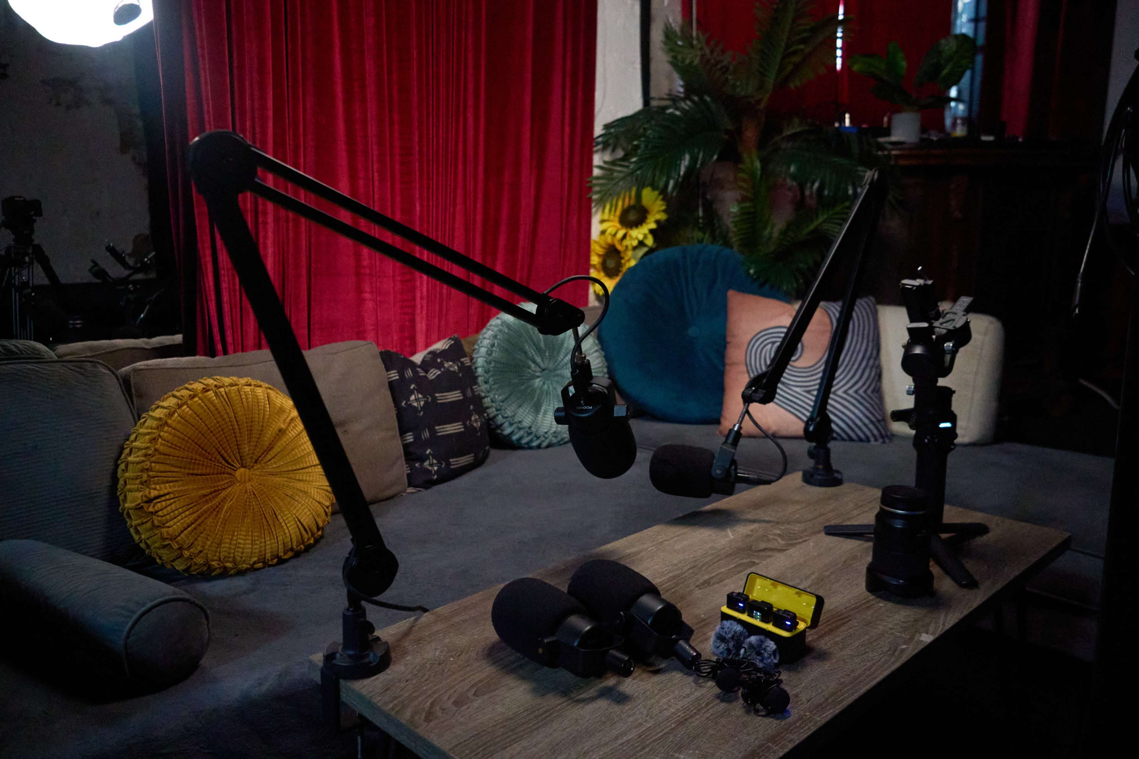 The image shows a podcast setup with multiple microphone stands, a wooden table, and a colorful couch adorned with pillows.