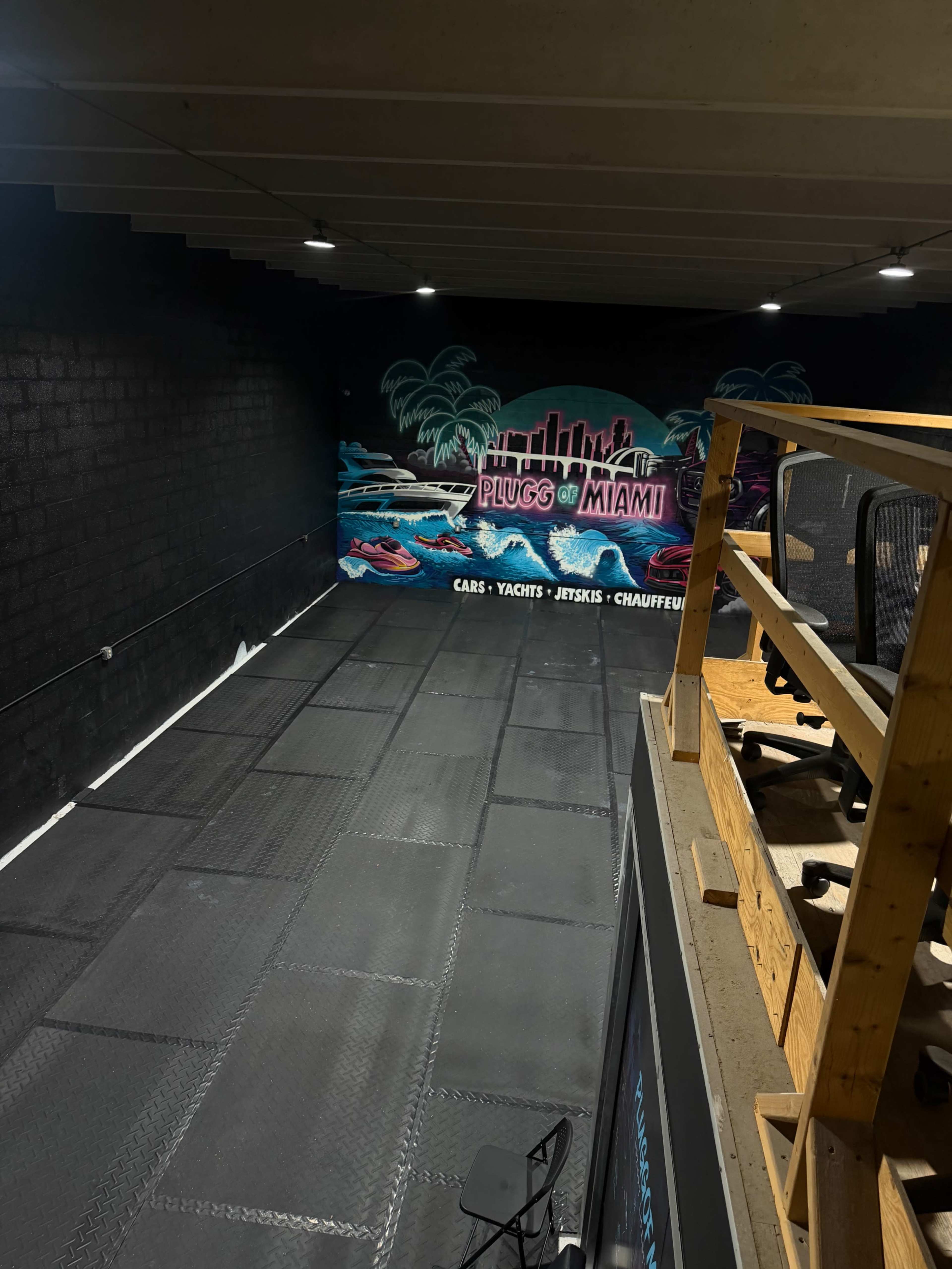 The image shows a dimly lit room with a black wall featuring a colorful mural of a Miami skyline, alongside a wooden structure and rubber flooring.