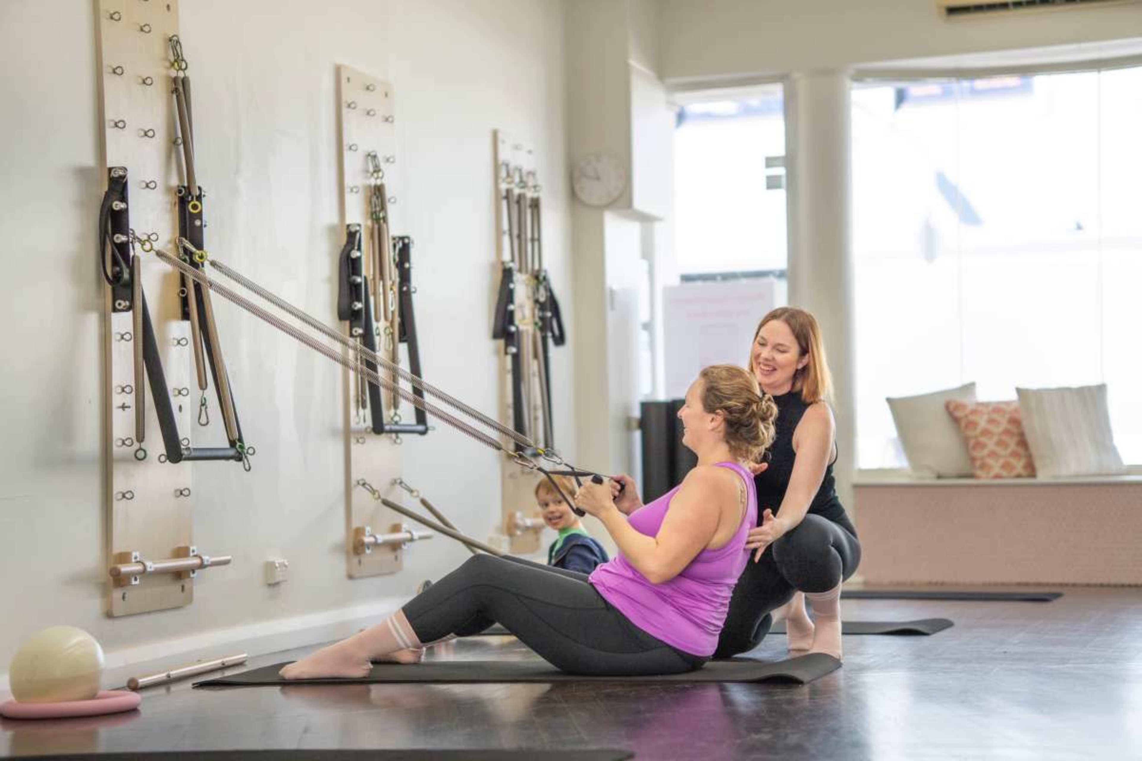 Two women are engaged in a Pilates workout using reformer equipment, while a child sits on the floor in the background.