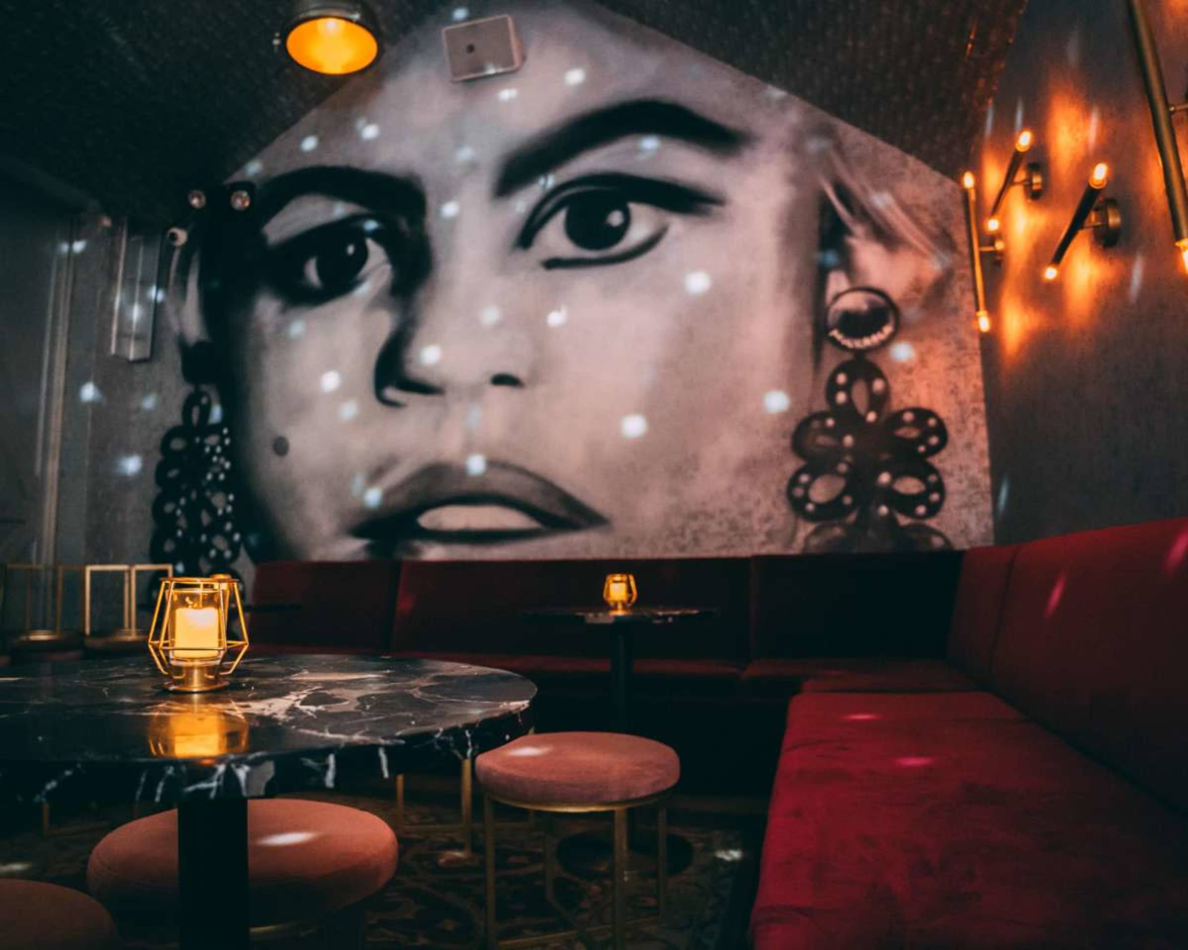 The image shows a dimly lit lounge with a large mural of a face on the wall, featuring gold accents and modern furniture.