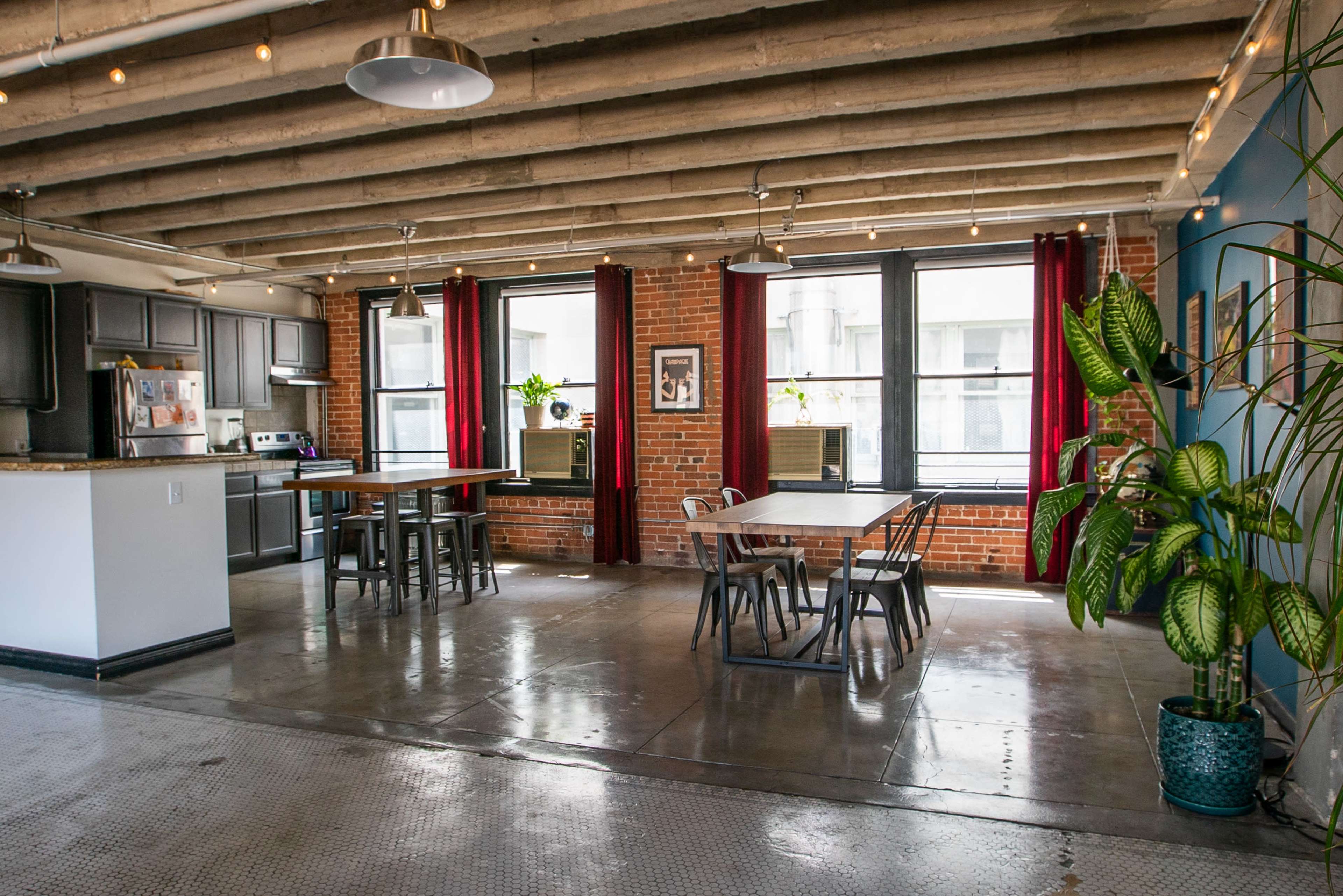 The image shows an open-concept kitchen and dining area with exposed brick walls, concrete floors, and large windows adorned with red curtains.