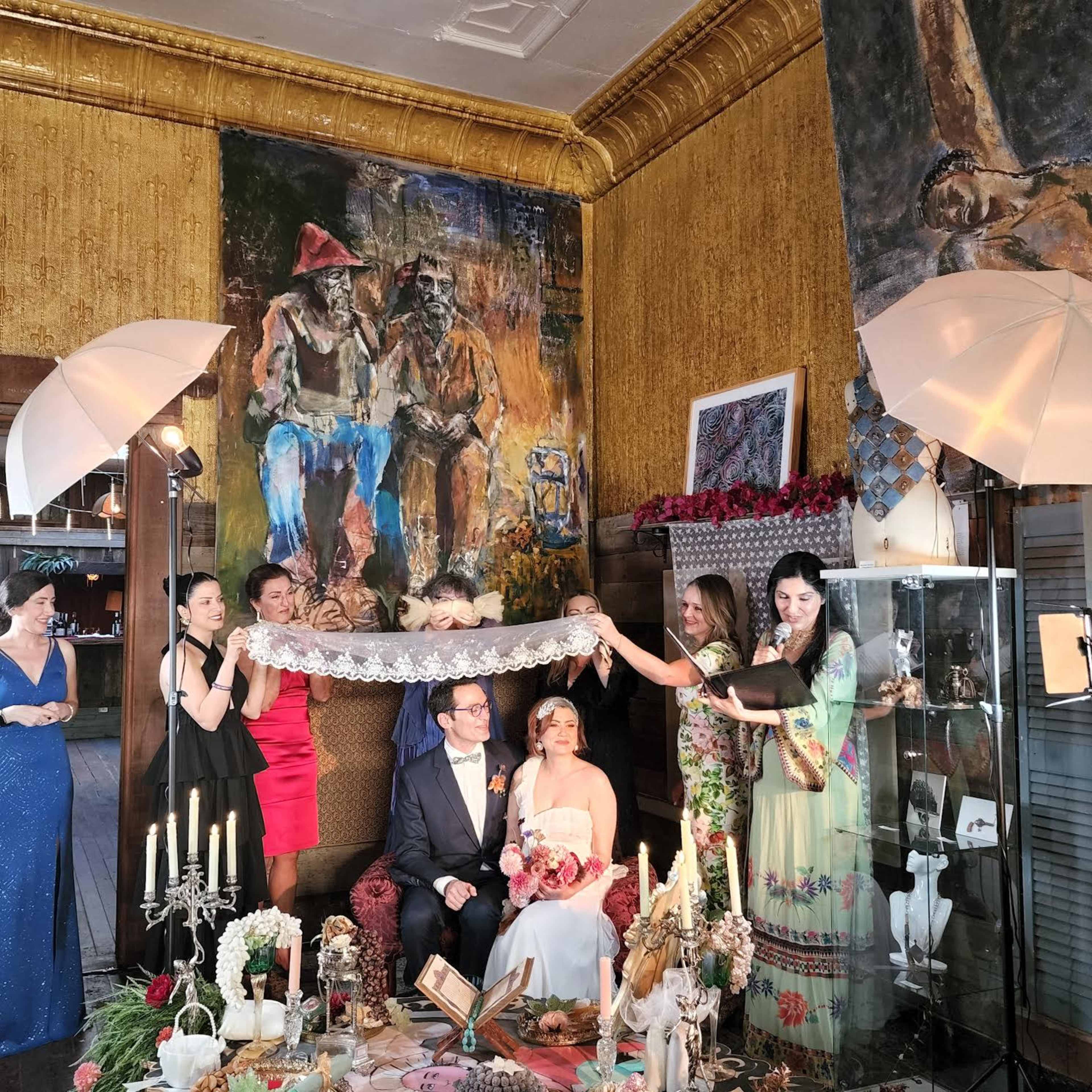 A bride and groom sit at the center of a decorated room, surrounded by guests holding a lace veil while colorful paintings adorn the walls.