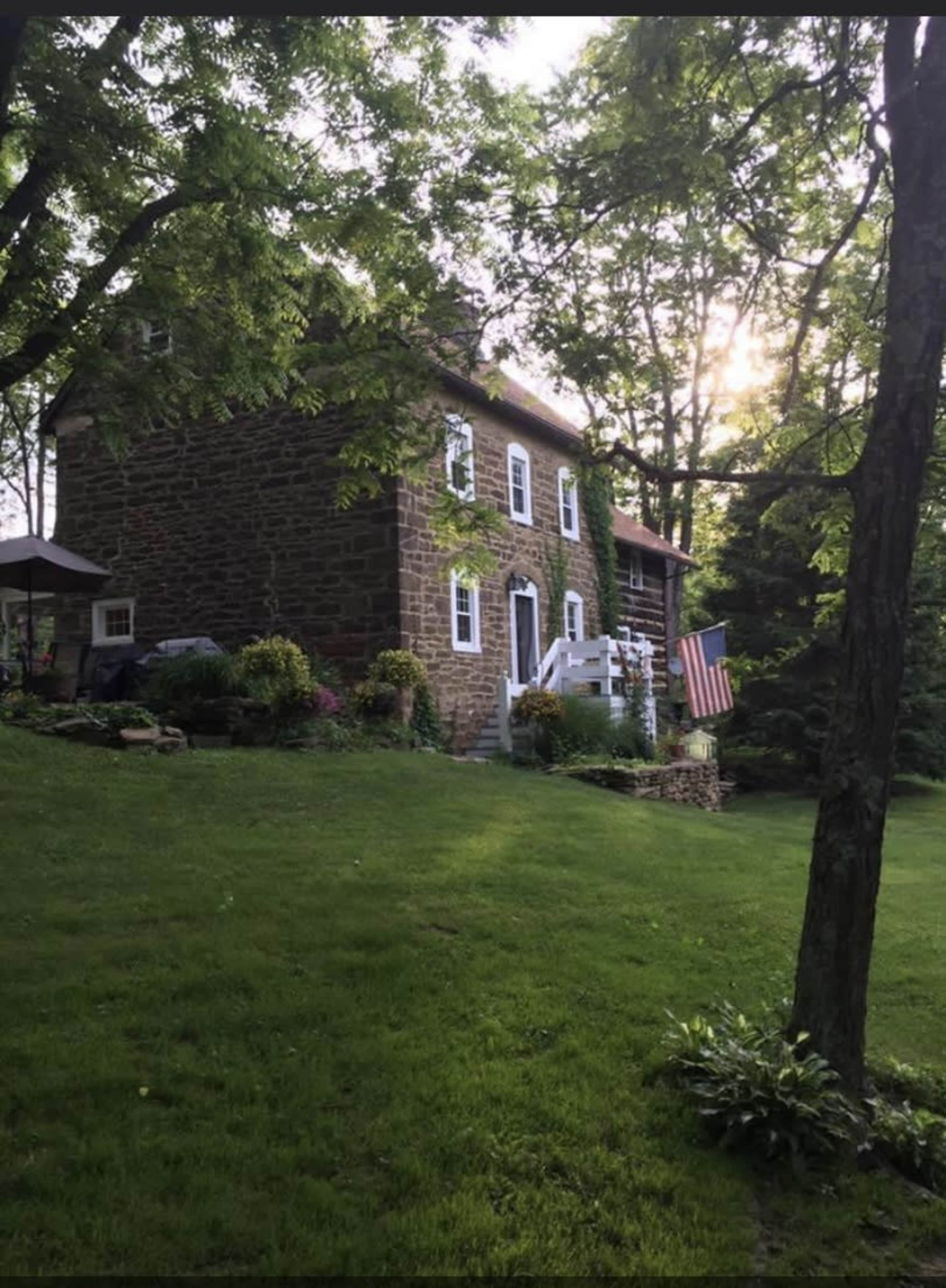 A two-story stone house is surrounded by green grass and trees, with a small American flag displayed on the porch.