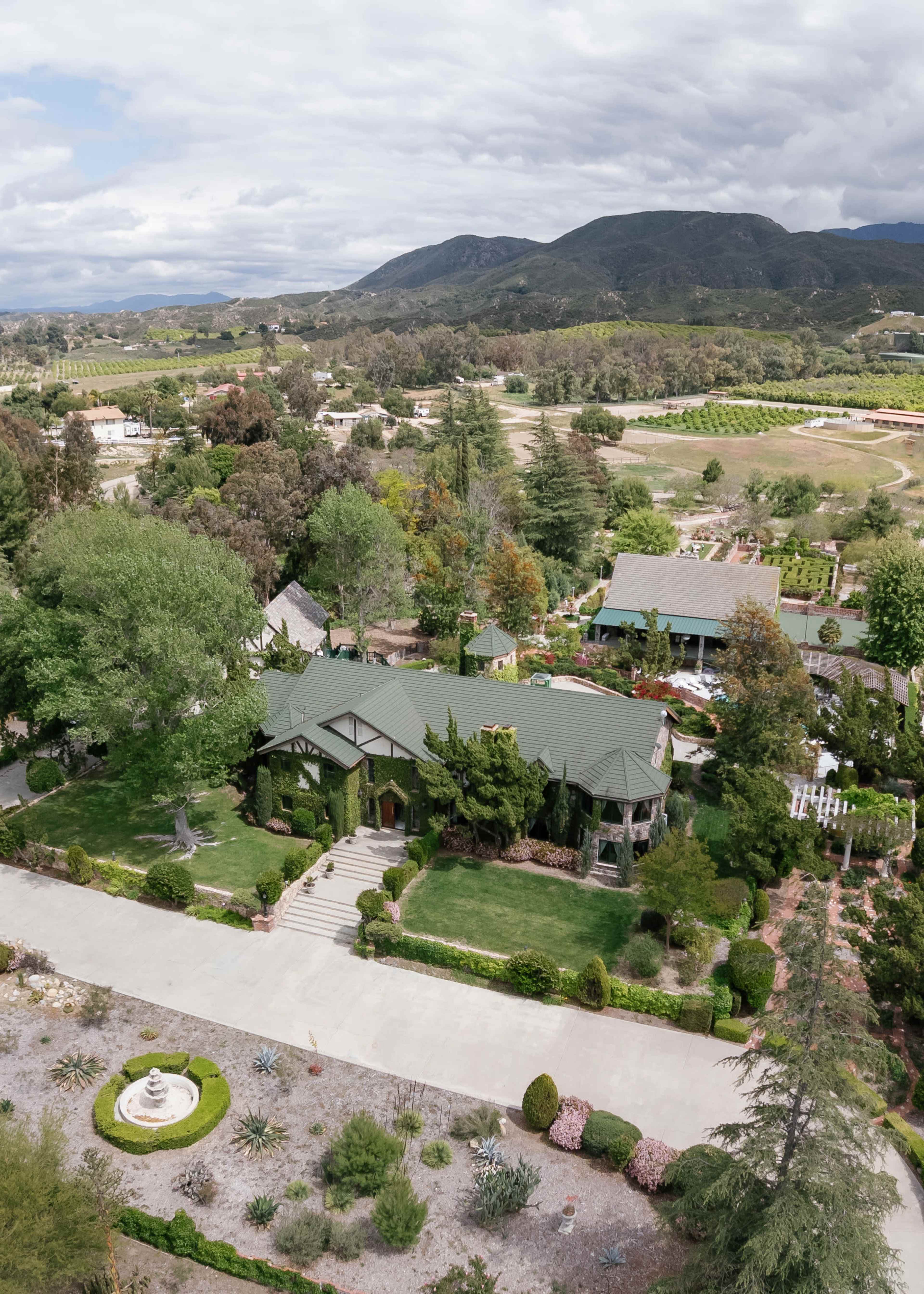 The image showcases a large, landscaped property with a sprawling green lawn, a distinctive house with a sloped roof, and surrounding gardens, set against a backdrop of rolling hills and mountains.