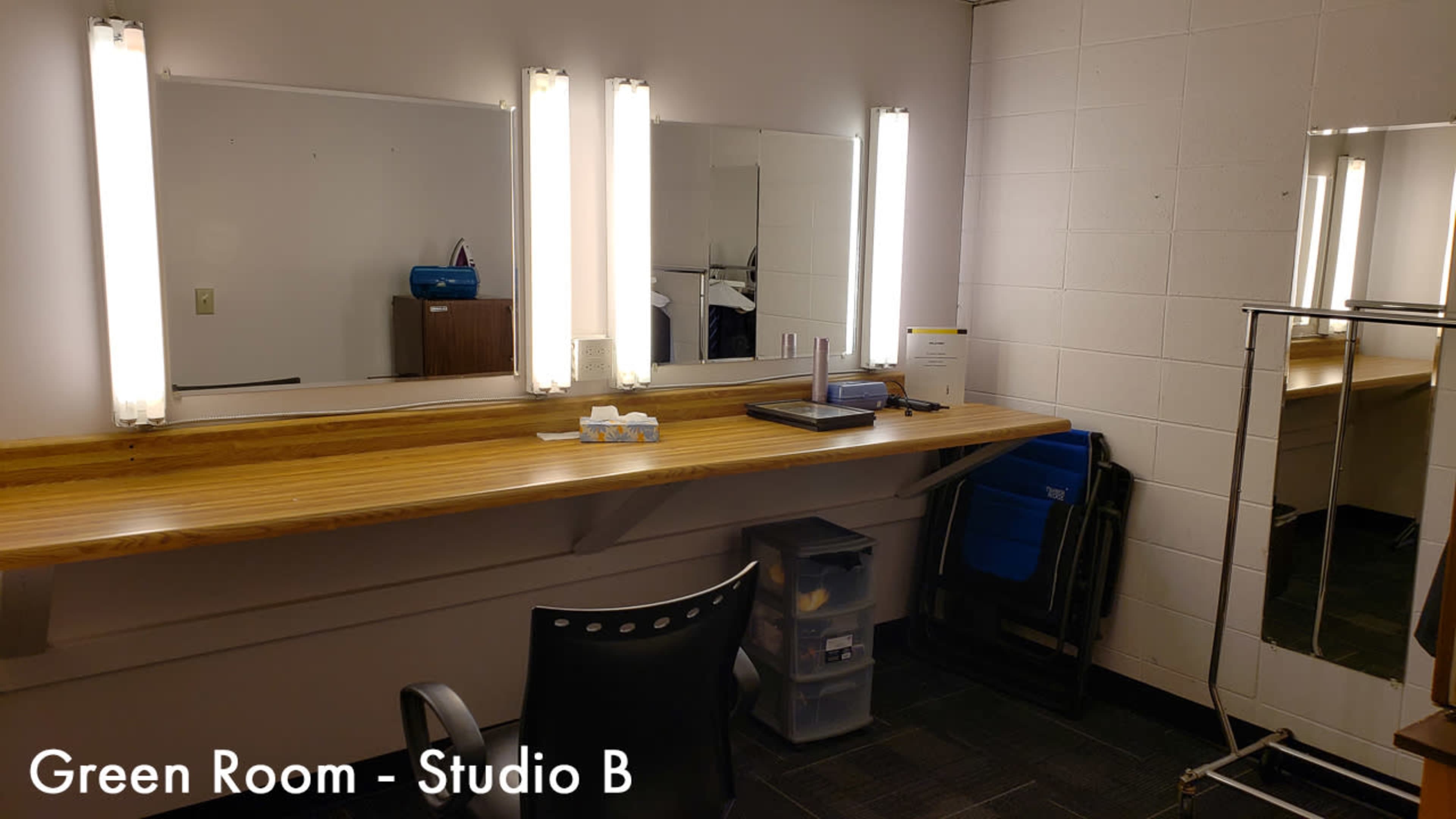 A green room with a long wooden table, three mirrors with bright lights, a chair, and some blue folding chairs against the wall.