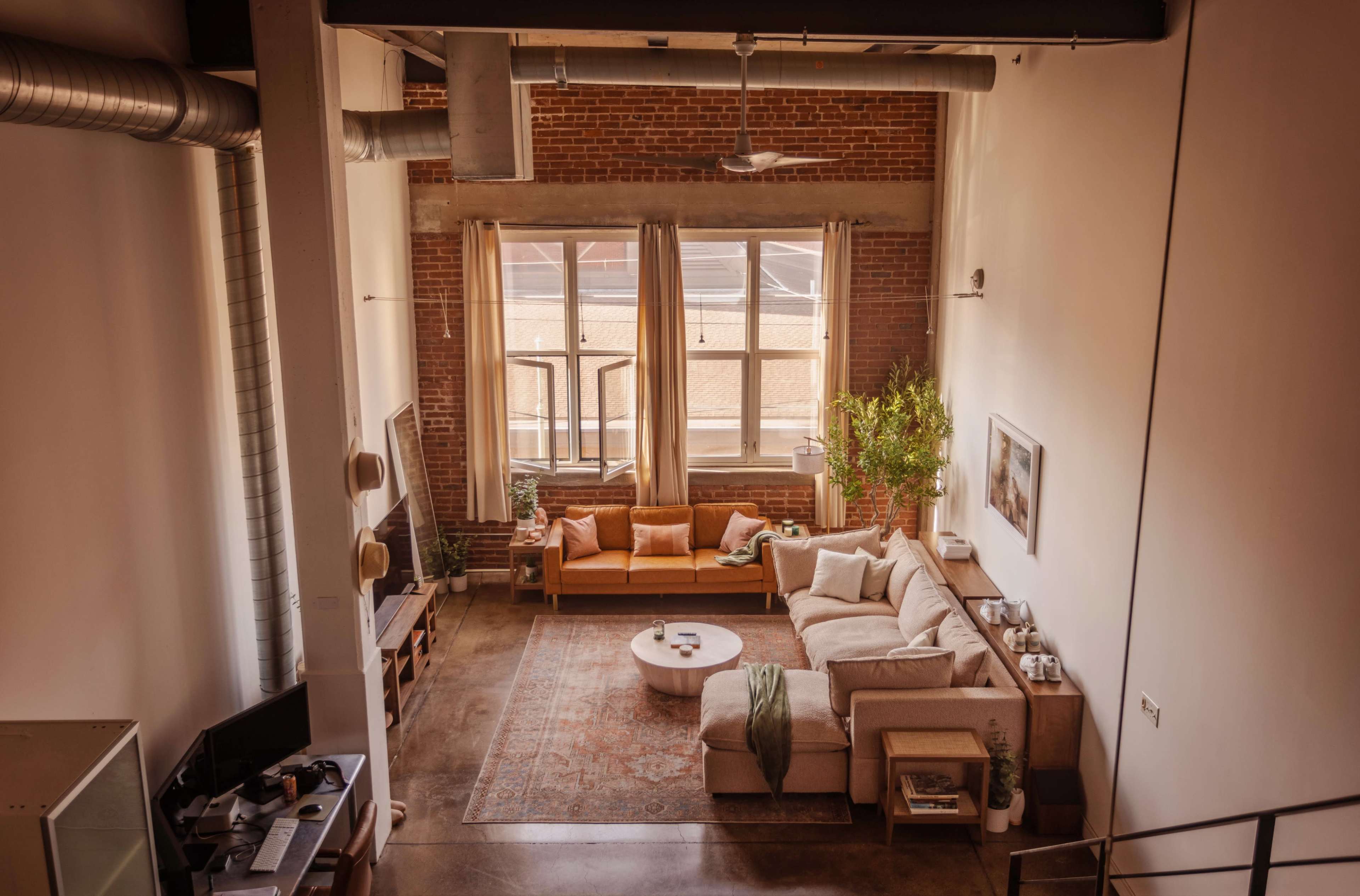 A spacious living room features large windows, exposed brick walls, a sectional sofa, and a coffee table, with plants and decorative elements adding to the décor.