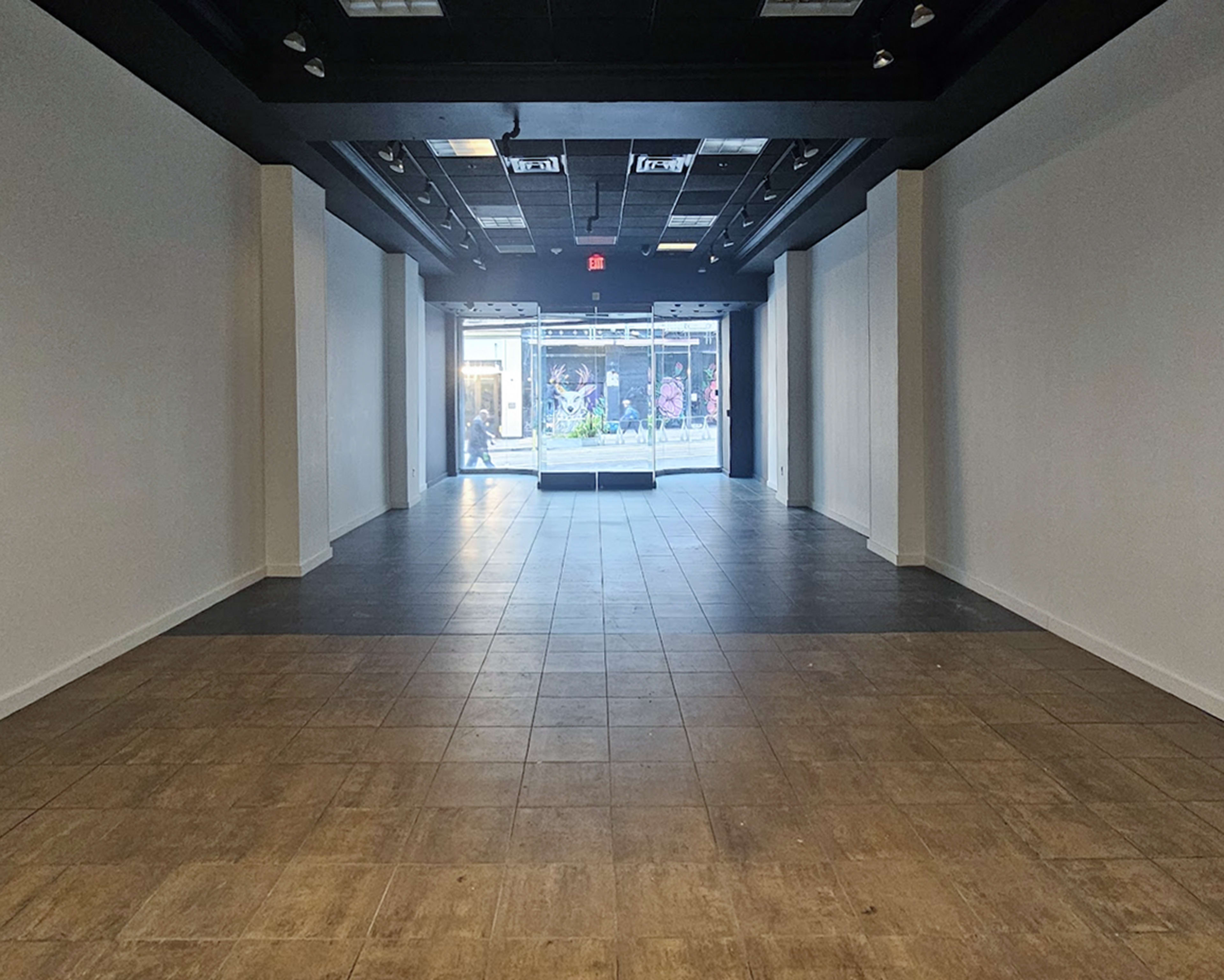The image shows an empty retail space with tiled flooring and large glass windows at the front, leading to a street outside.