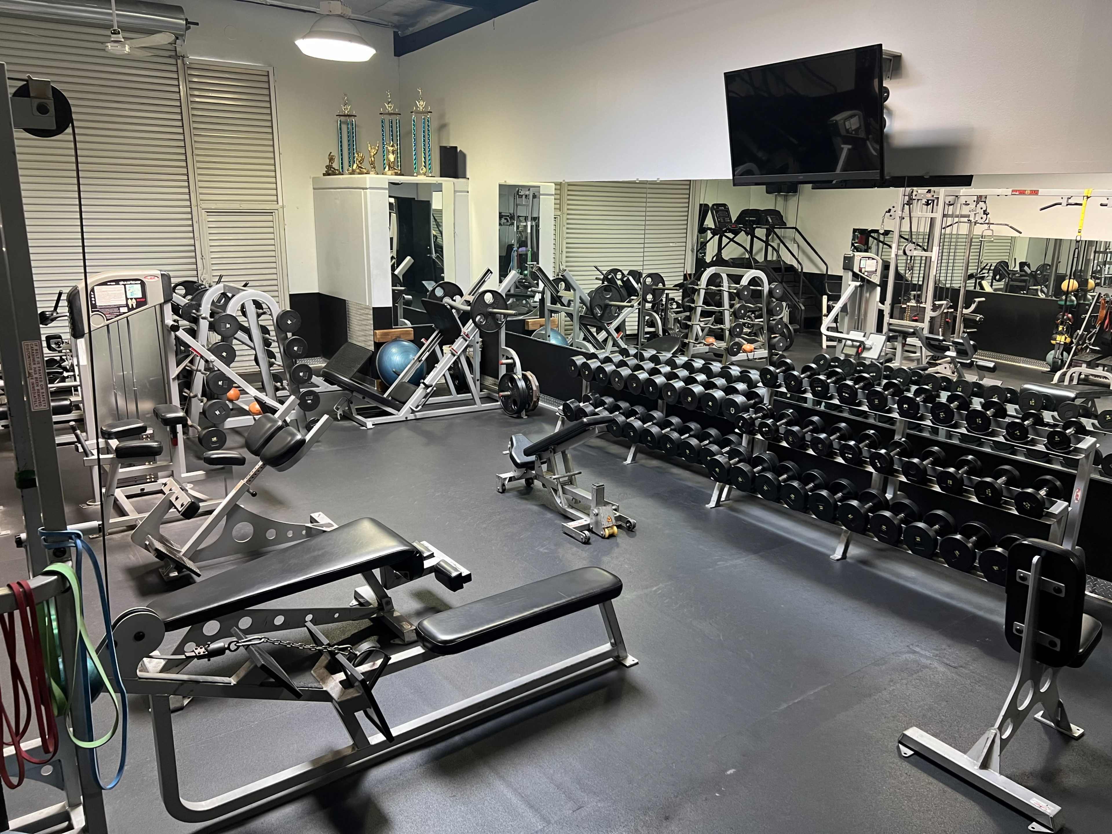 The image shows a gym interior with various workout equipment, including weight benches, dumbbells, and a television mounted on the wall.