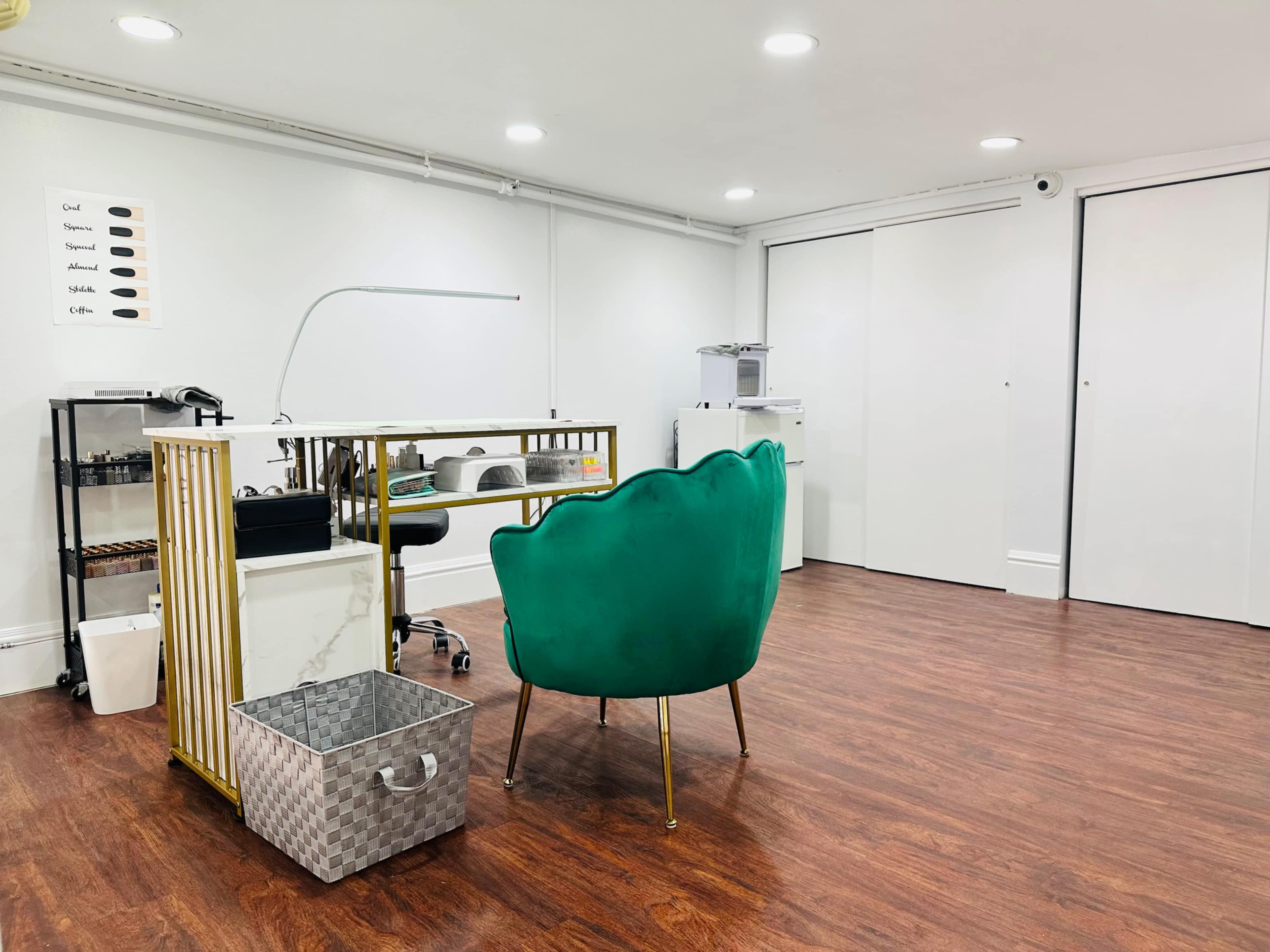 The image shows a minimalist nail salon with a green velvet chair, a manicure table, and a storage cart against a light-colored wall.