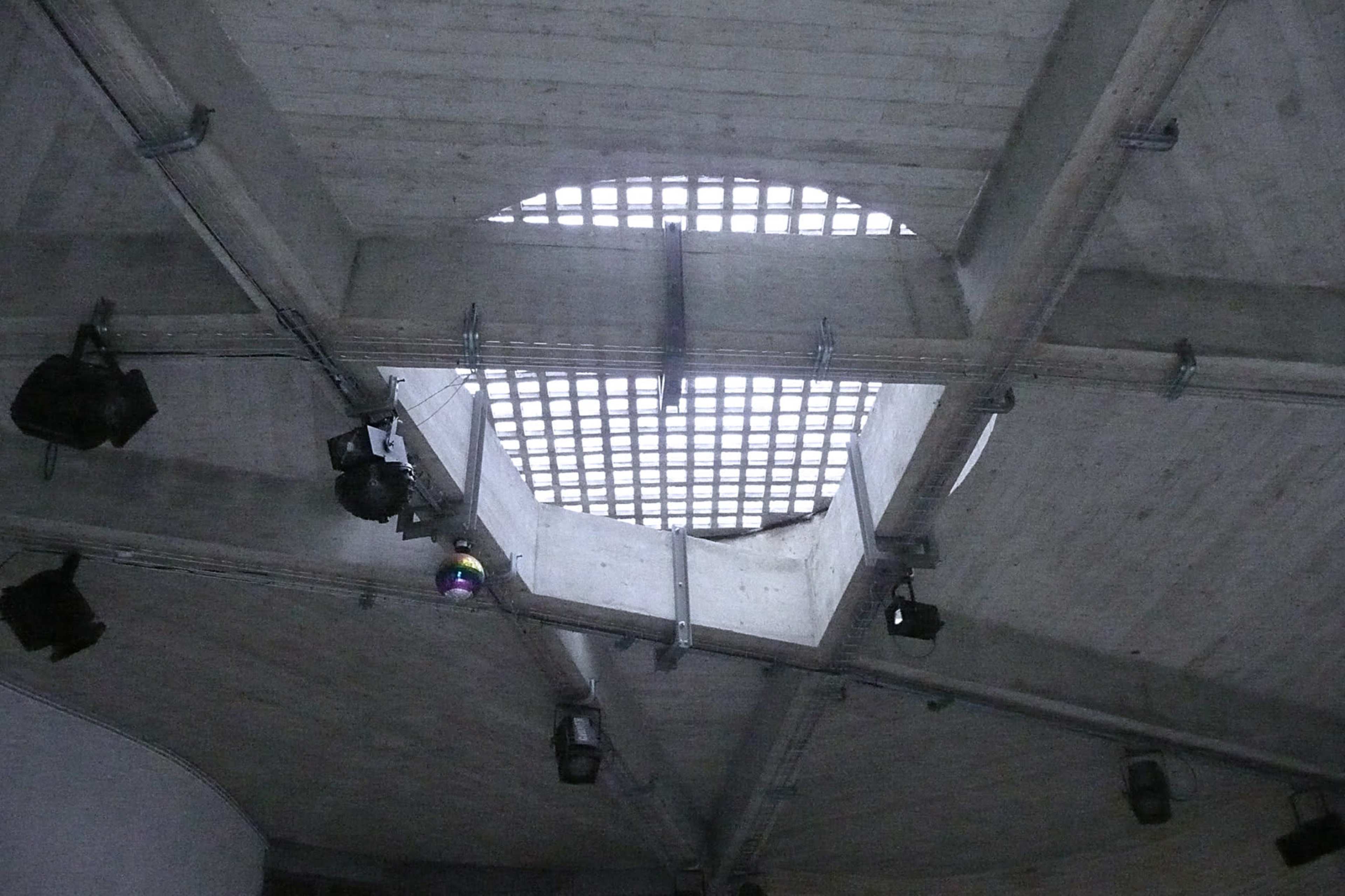 A portion of a concrete ceiling with a square opening letting light in, surrounded by structural beams and spotlights.