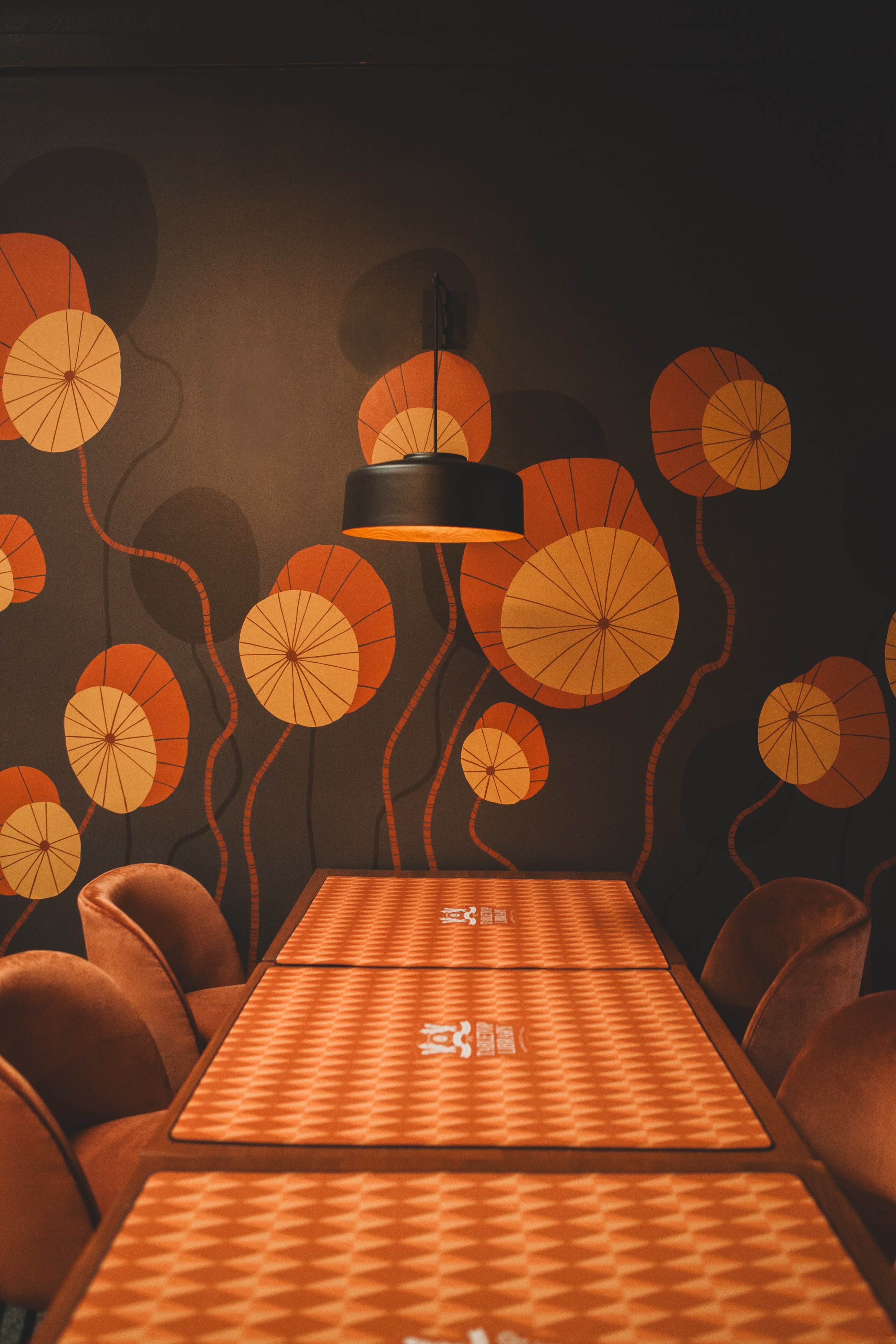 The image shows a dimly lit dining area with brown walls featuring abstract floral patterns and tables covered with orange textured tablecloths.