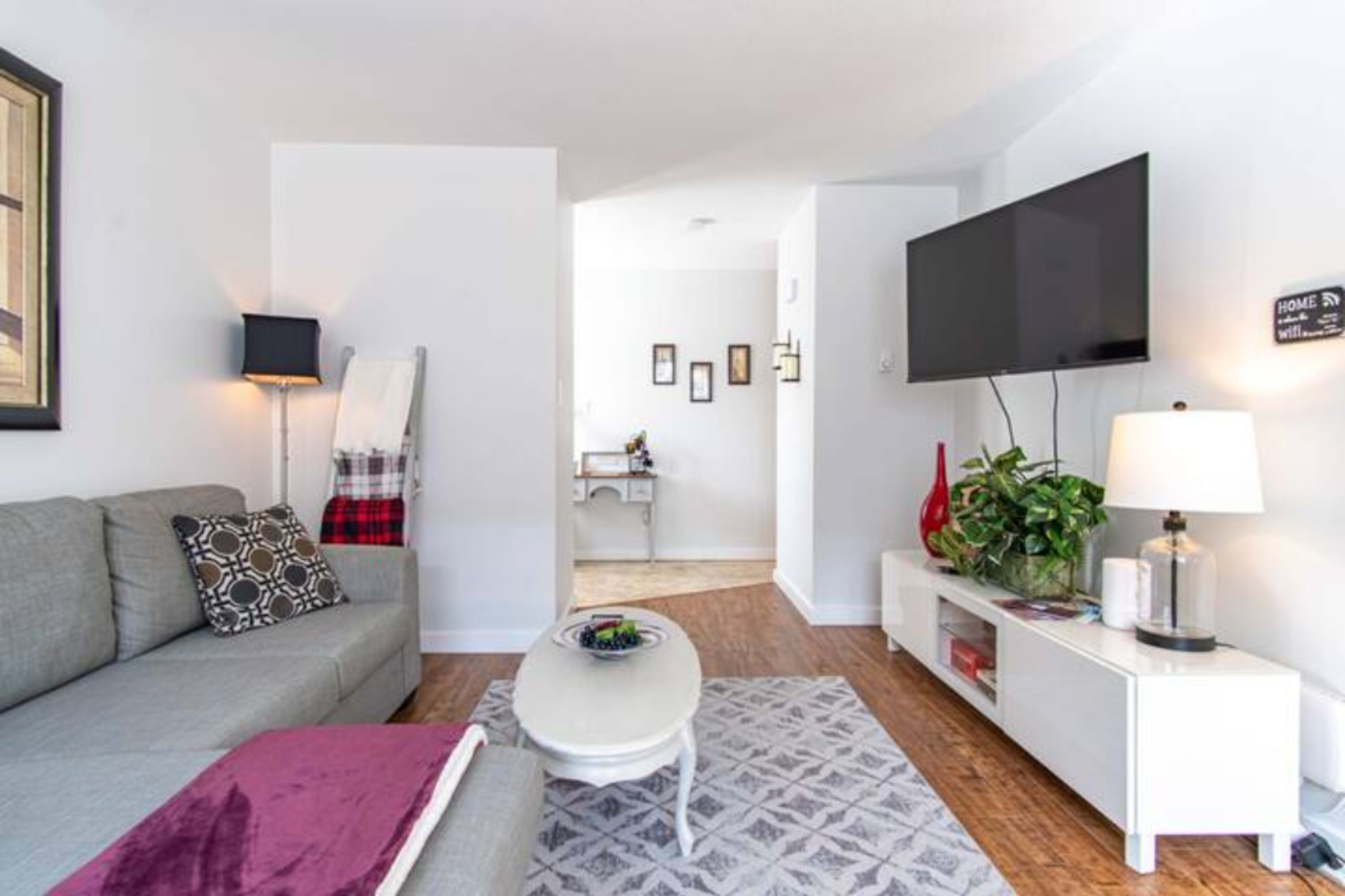 A modern living room features a gray couch, a round coffee table, and a wall-mounted television, with a view into an adjoining room that has a desk and decorative items.