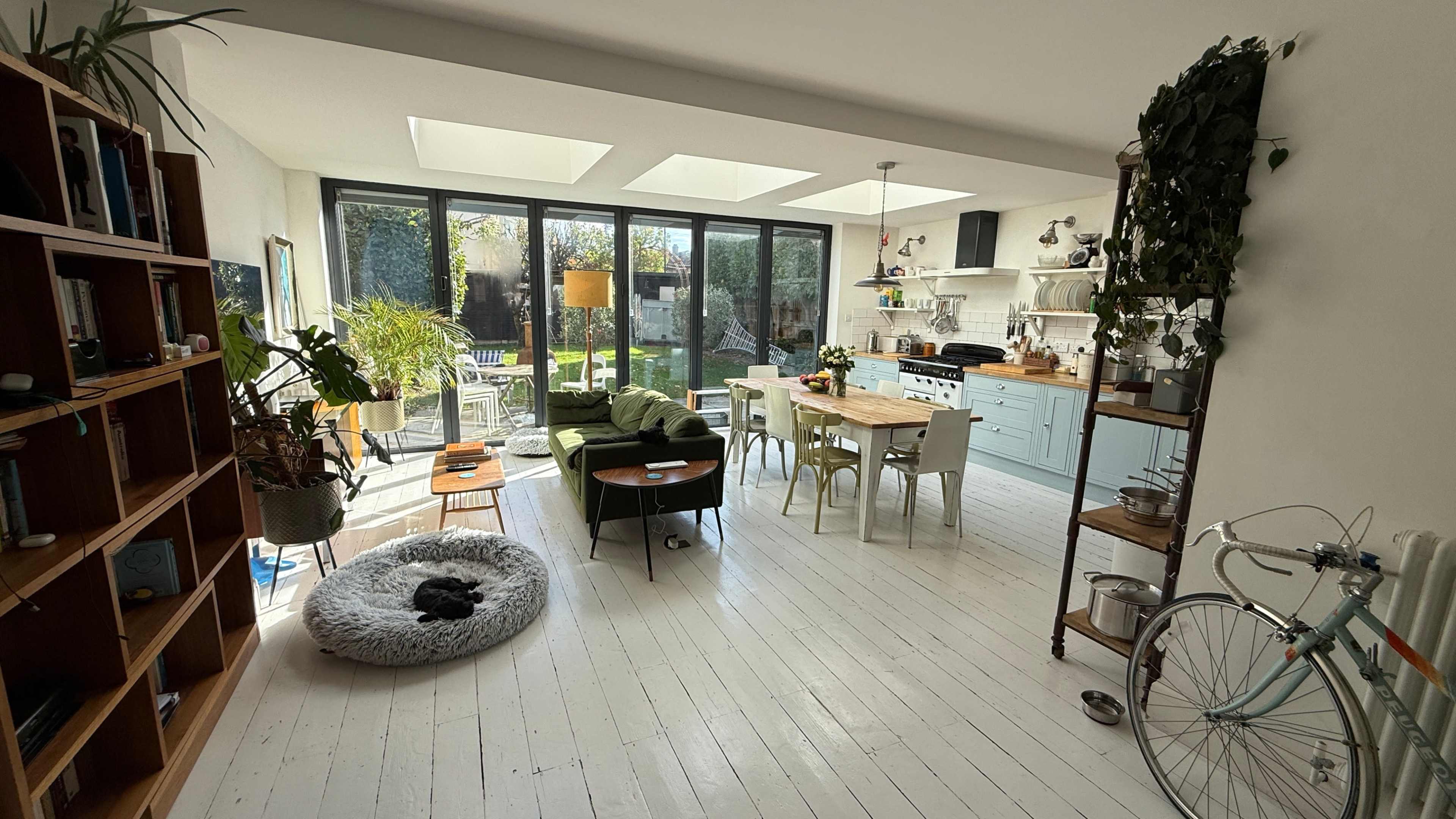 A modern, well-lit open-plan living area features a sitting space with green chairs, a dining table, and a kitchen with pastel-colored cabinetry, alongside a garden visible through large glass windows.