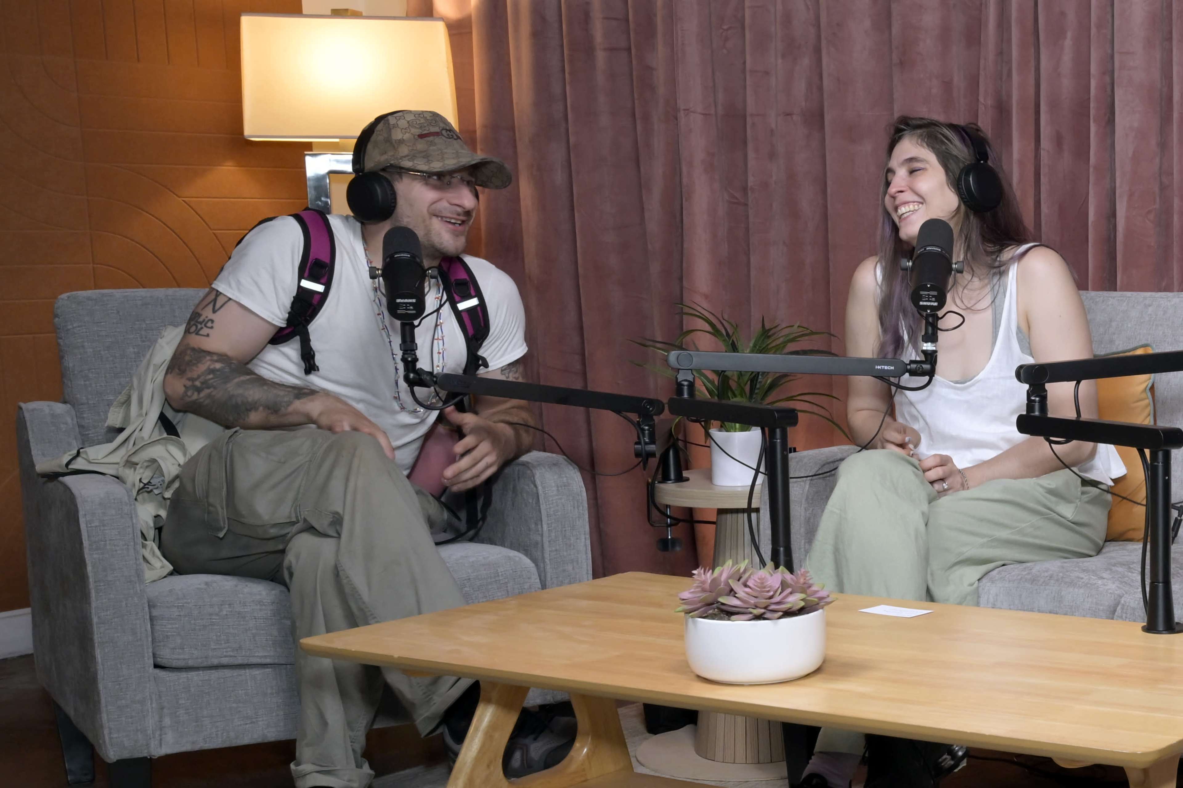 Two people sit on gray chairs in a studio setting, engaged in conversation while using microphones.