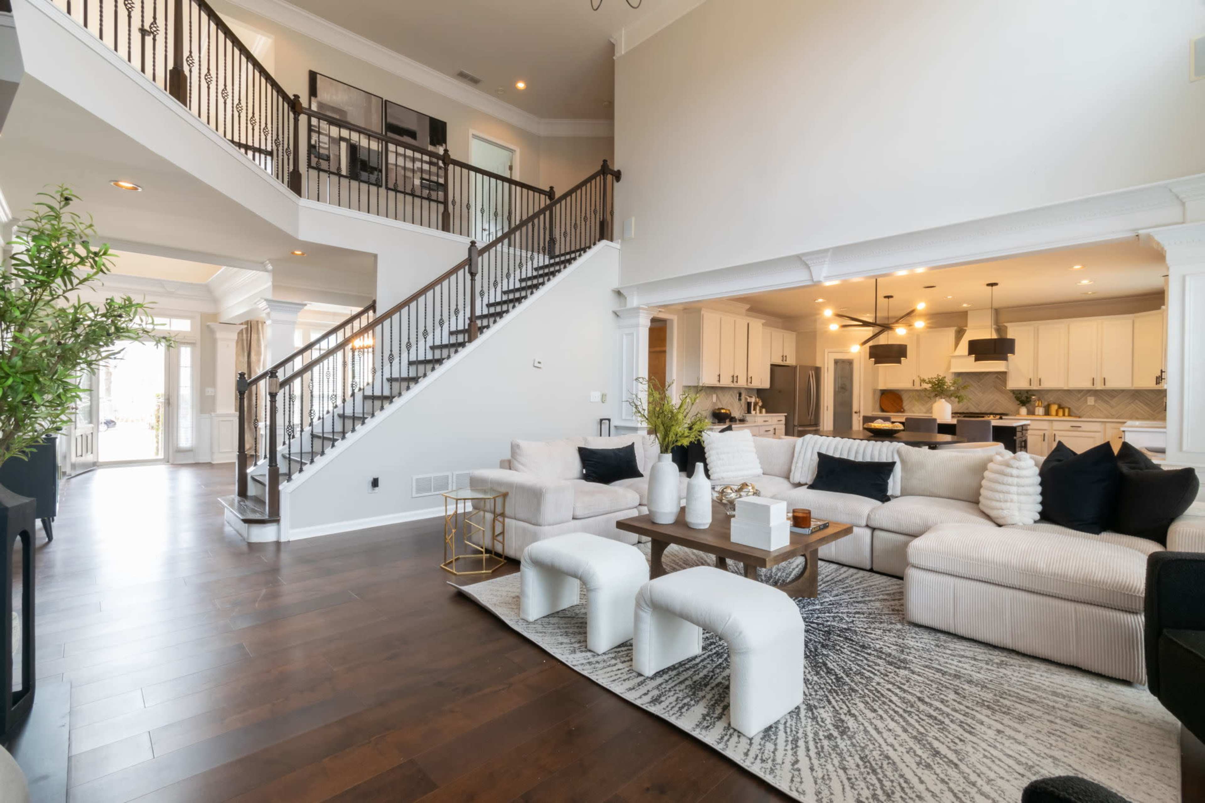 The image shows a spacious living room with a large sectional sofa, a coffee table, and a staircase leading to an upper level, adjacent to an open kitchen area.