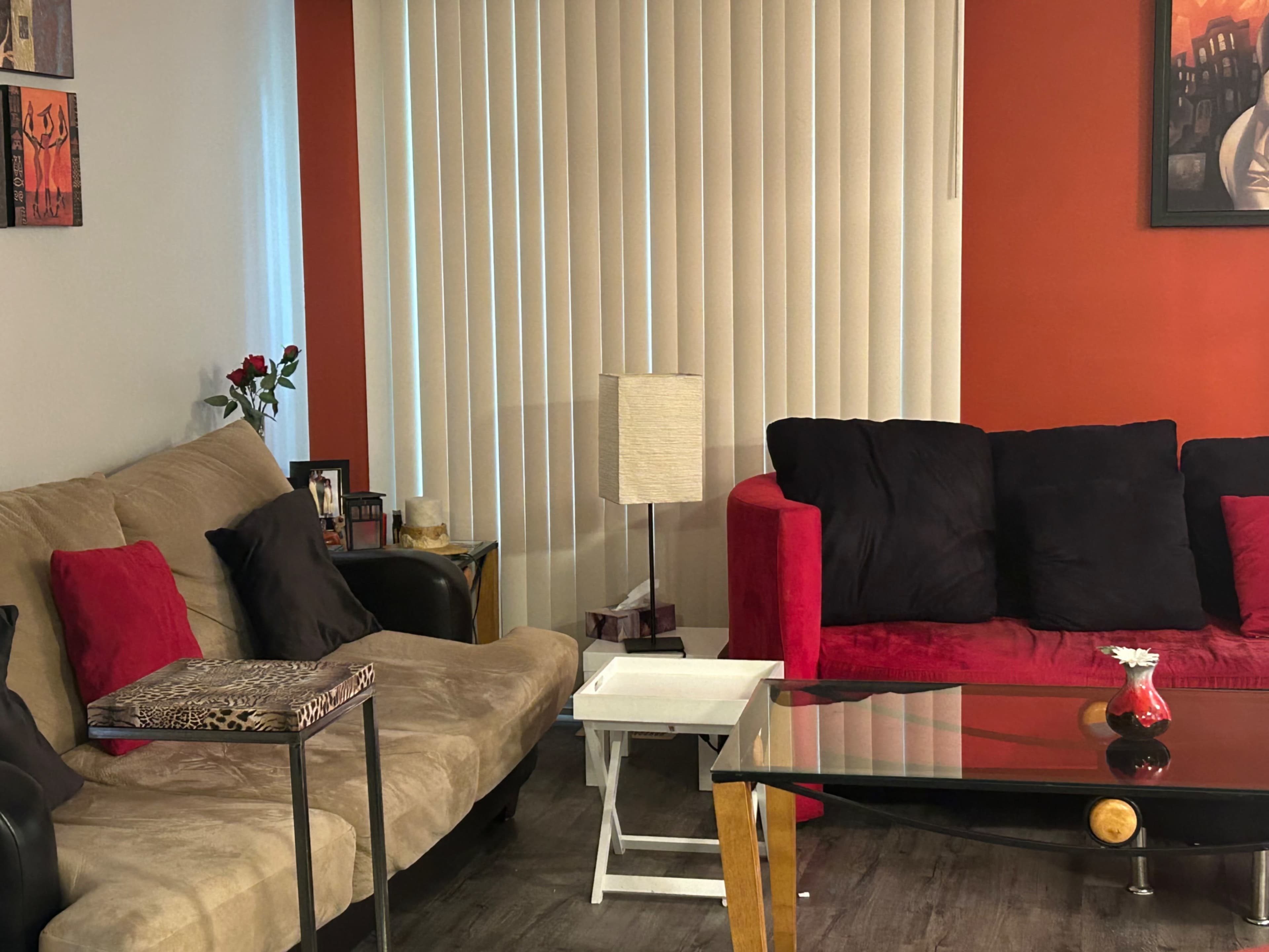The living room features a beige sofa, a red couch, a glass coffee table, and a lamp, with vertical blinds covering a window.