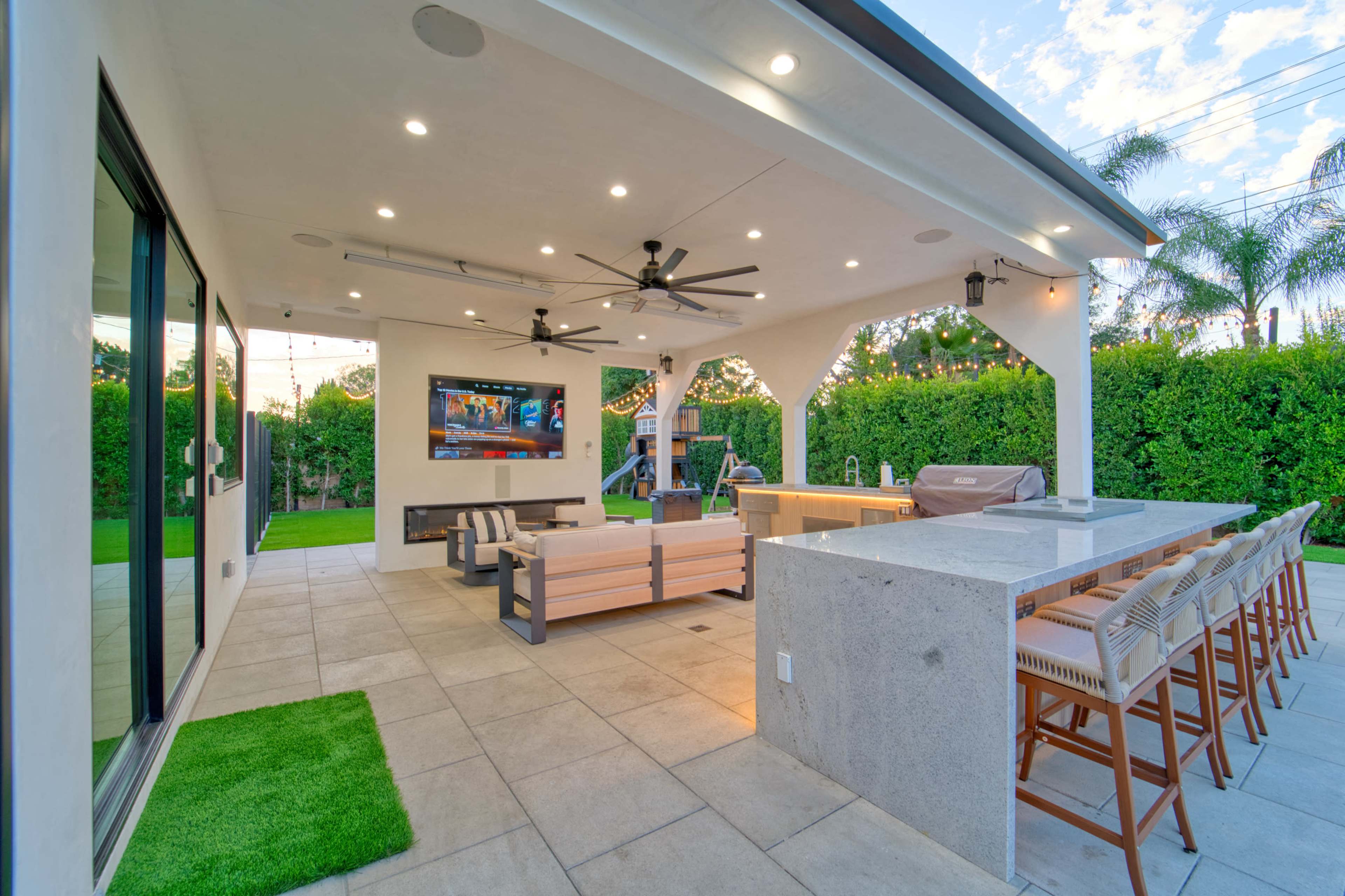 The image shows a modern outdoor patio with a kitchen area, seating, and a television, surrounded by lush greenery.