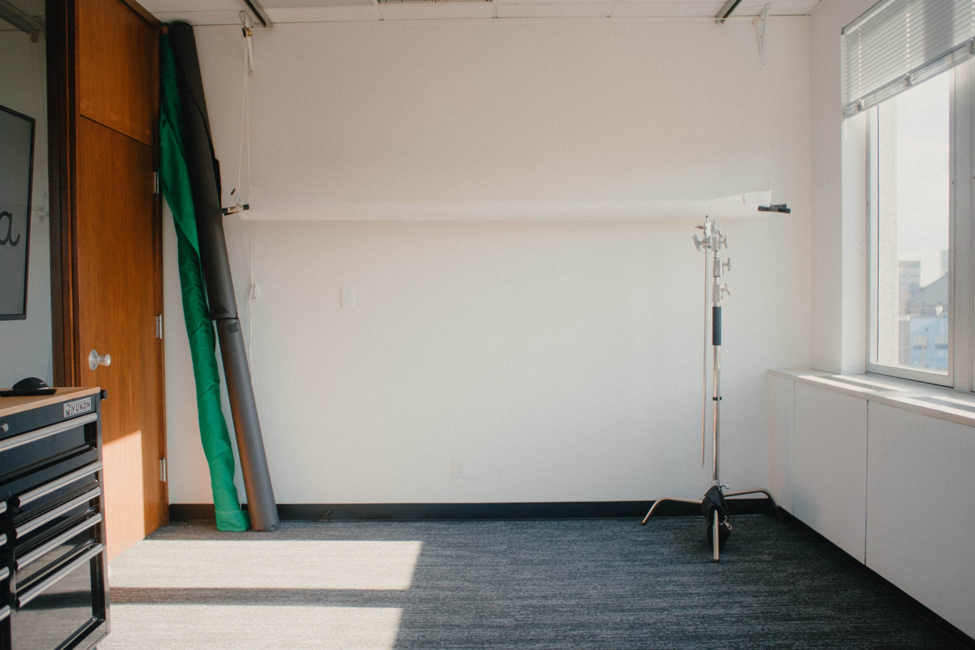 The image shows a bright, empty room with a green backdrop leaning against the wall and a light stand positioned near a window.