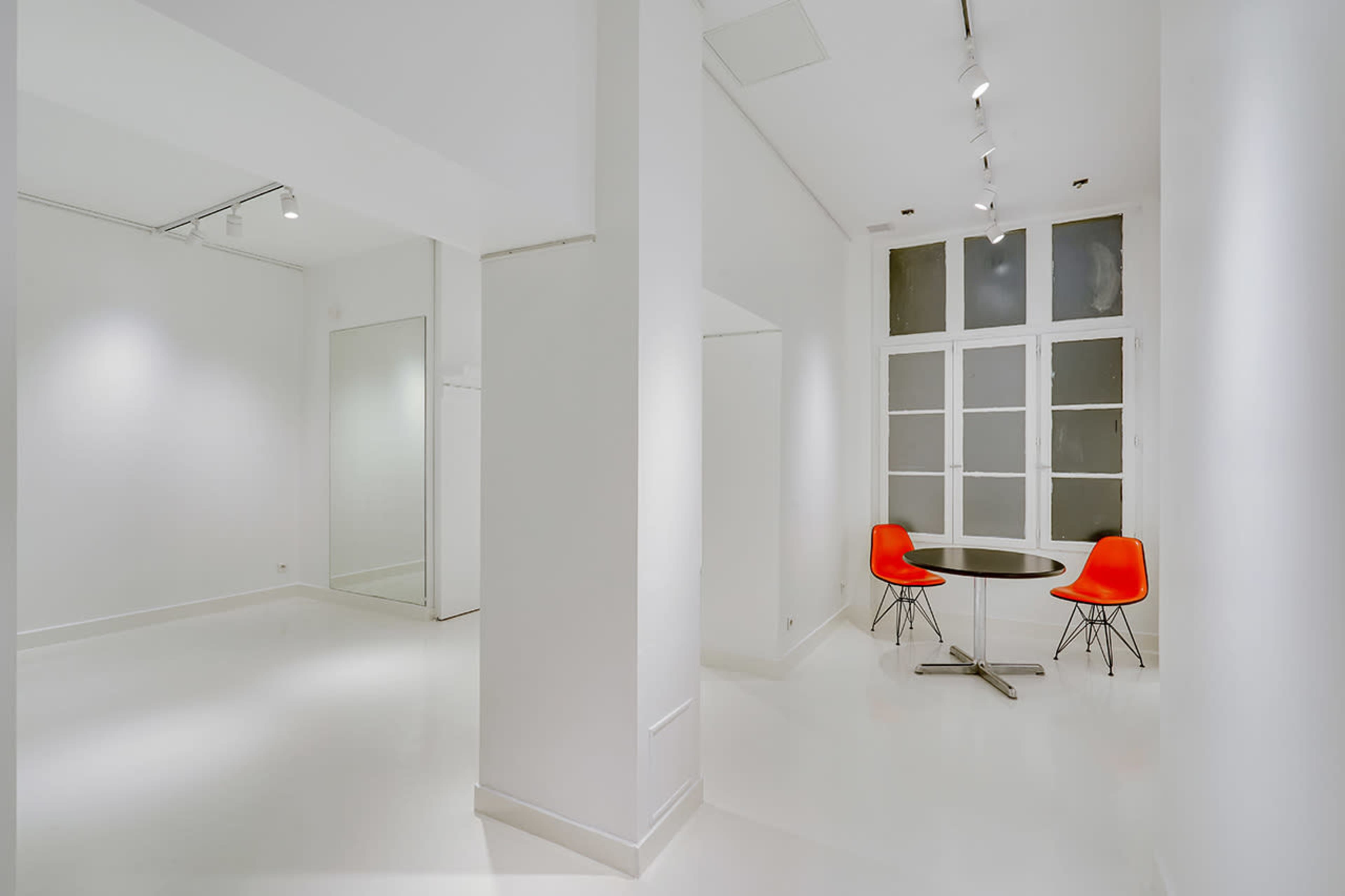 The image shows a minimalist, white-walled room featuring a round black table with two red chairs and a large window with multiple panes.