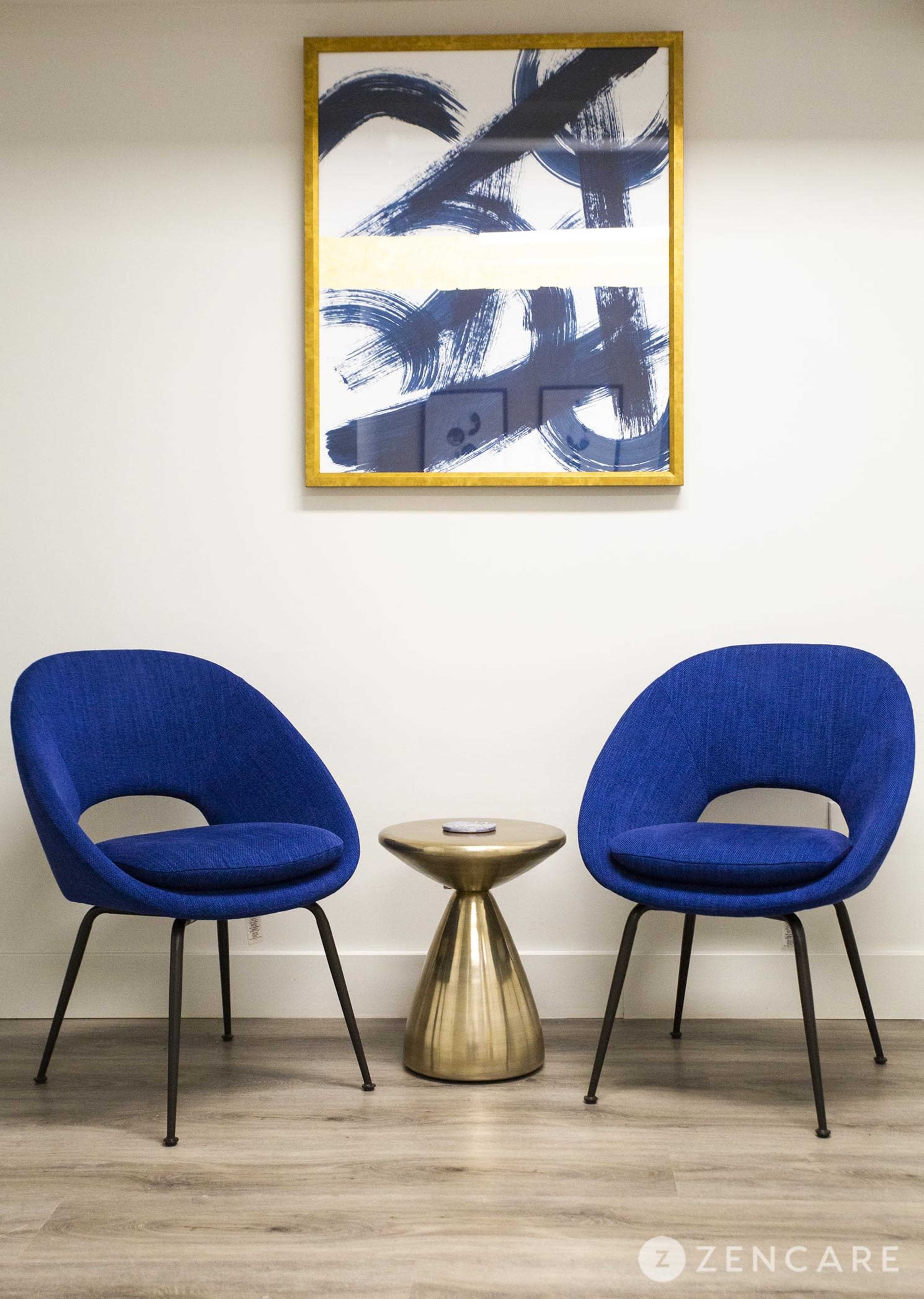 The image features two blue chairs facing each other across a small, gold accent table, with a large abstract painting hanging on the wall above them.