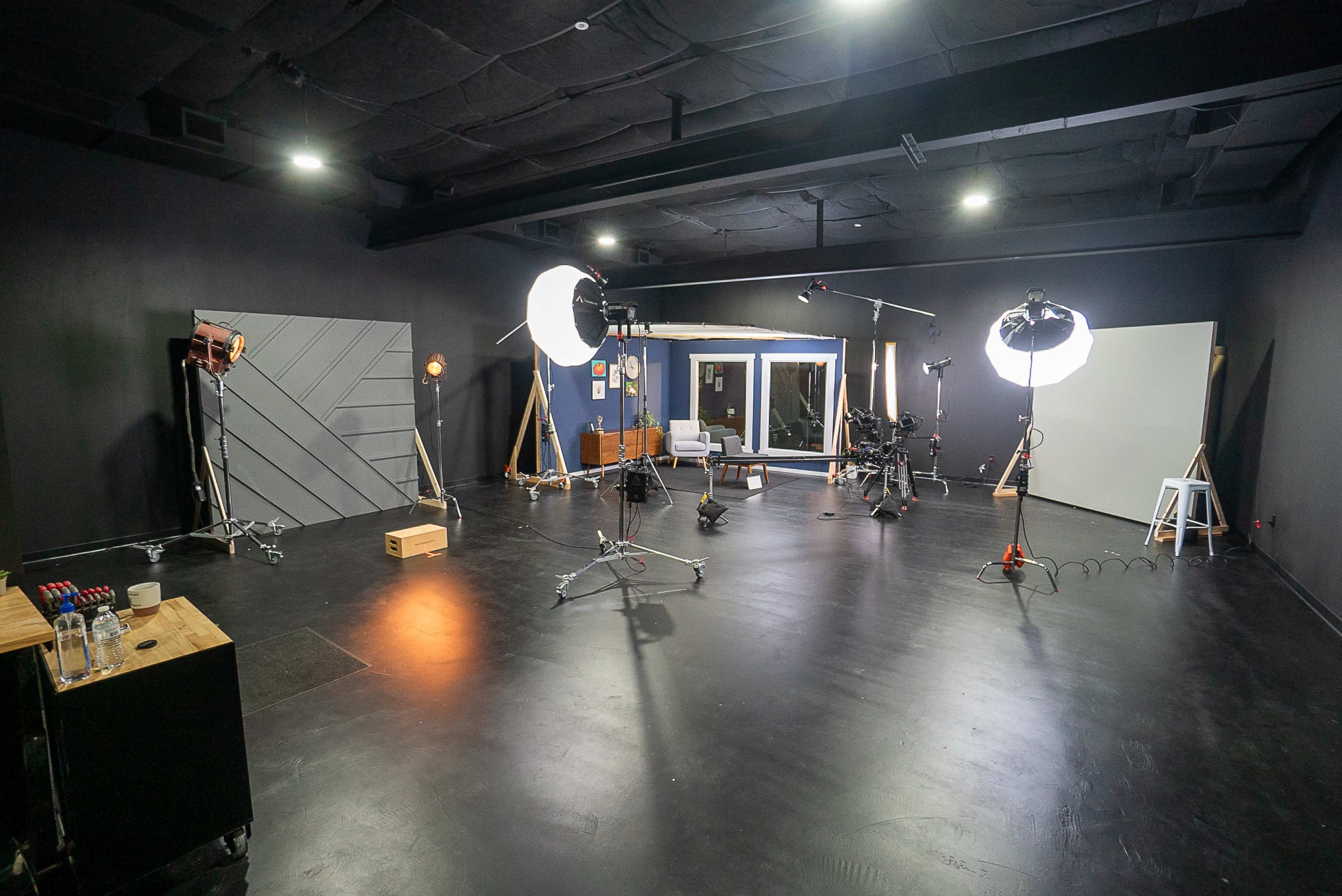 A dimly lit photography studio features various lighting equipment, a camera on a tripod, and a white backdrop.