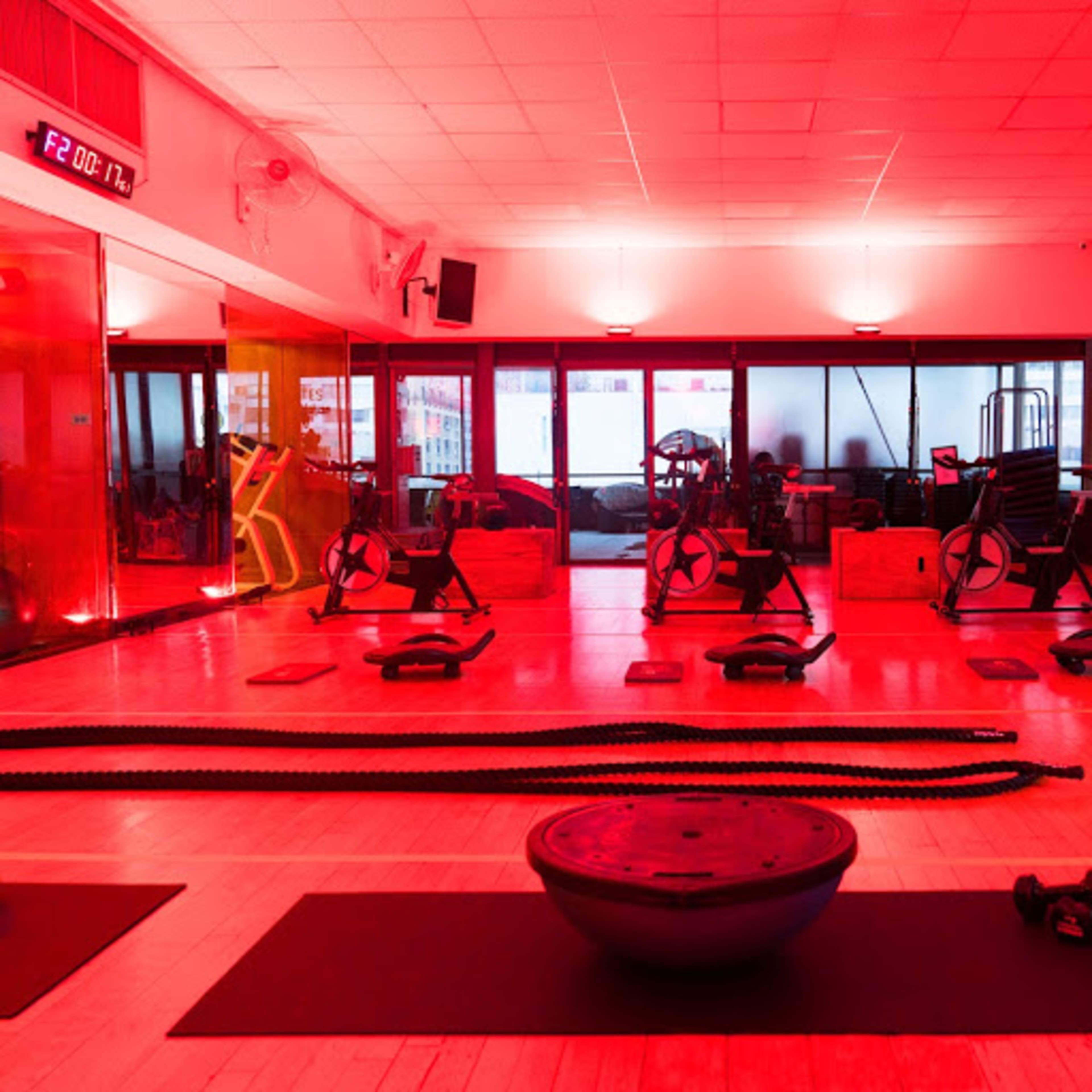 A gym interior with red lighting, featuring exercise equipment such as rowing machines, a stability ball, and battle ropes arranged on wooden flooring.
