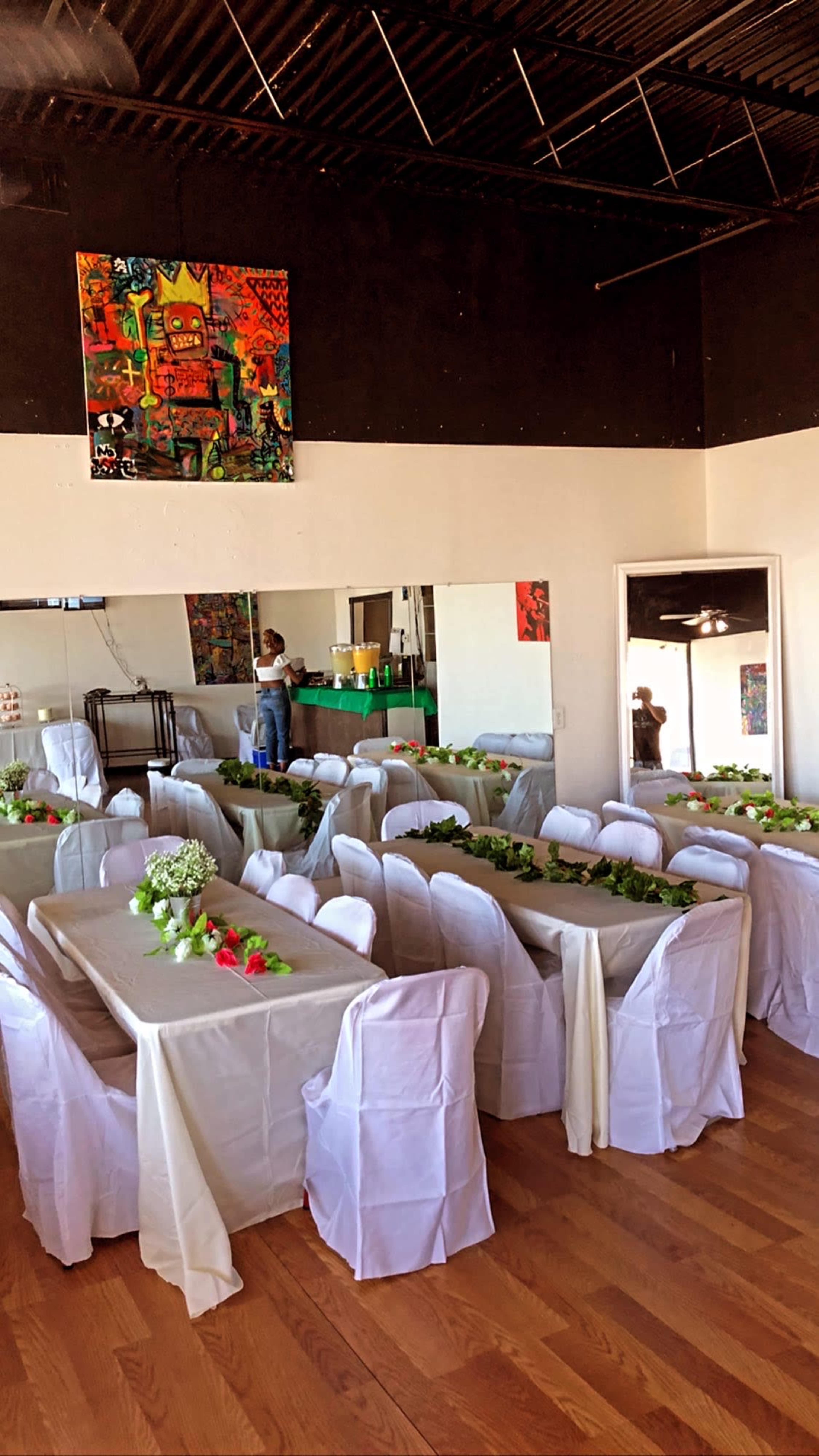 A dining area with multiple tables covered with white tablecloths and decorated with greenery is set up for an event.