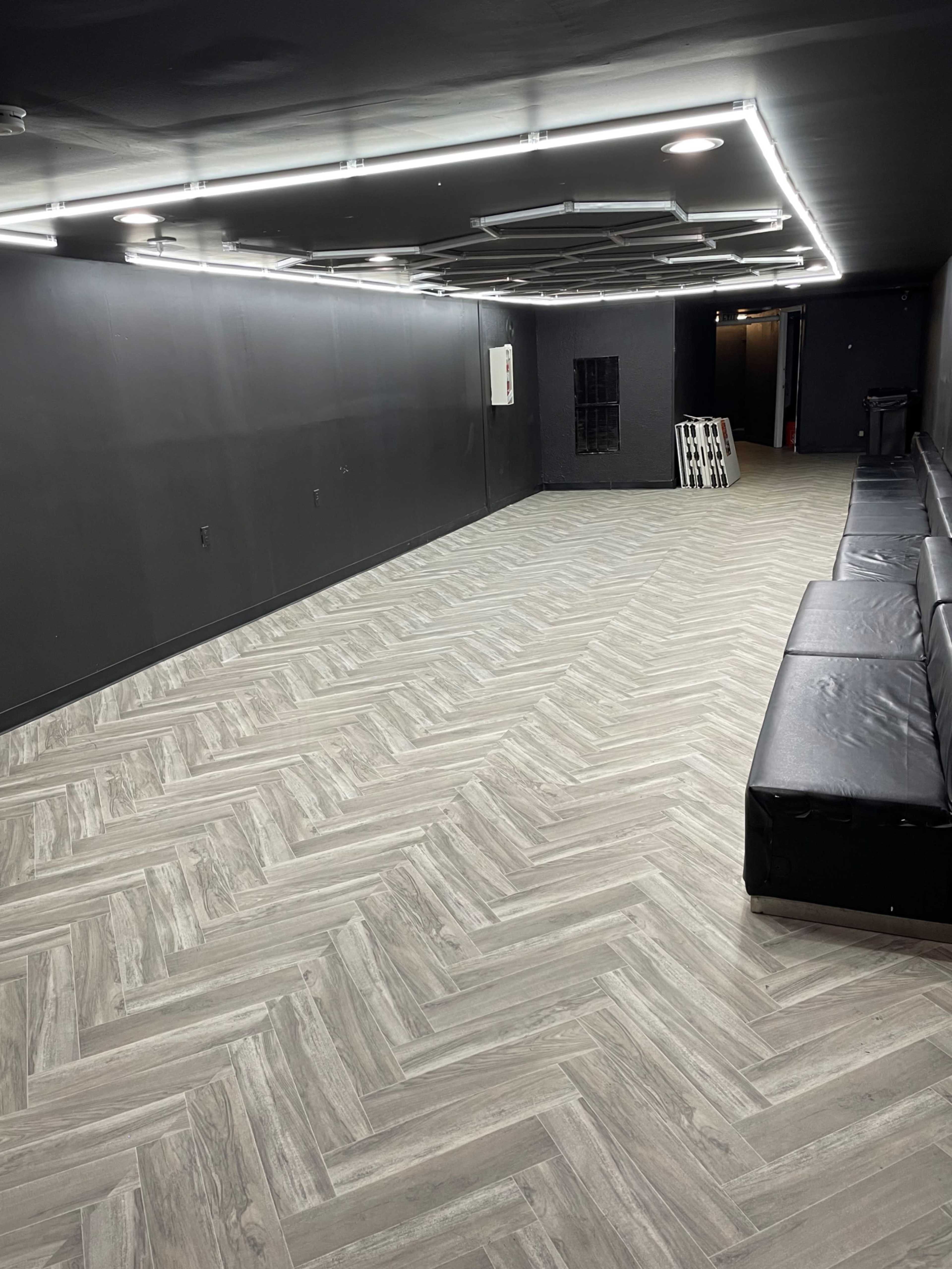 A spacious, empty room with dark walls, a herringbone-patterned wooden floor, and black seating along one side.