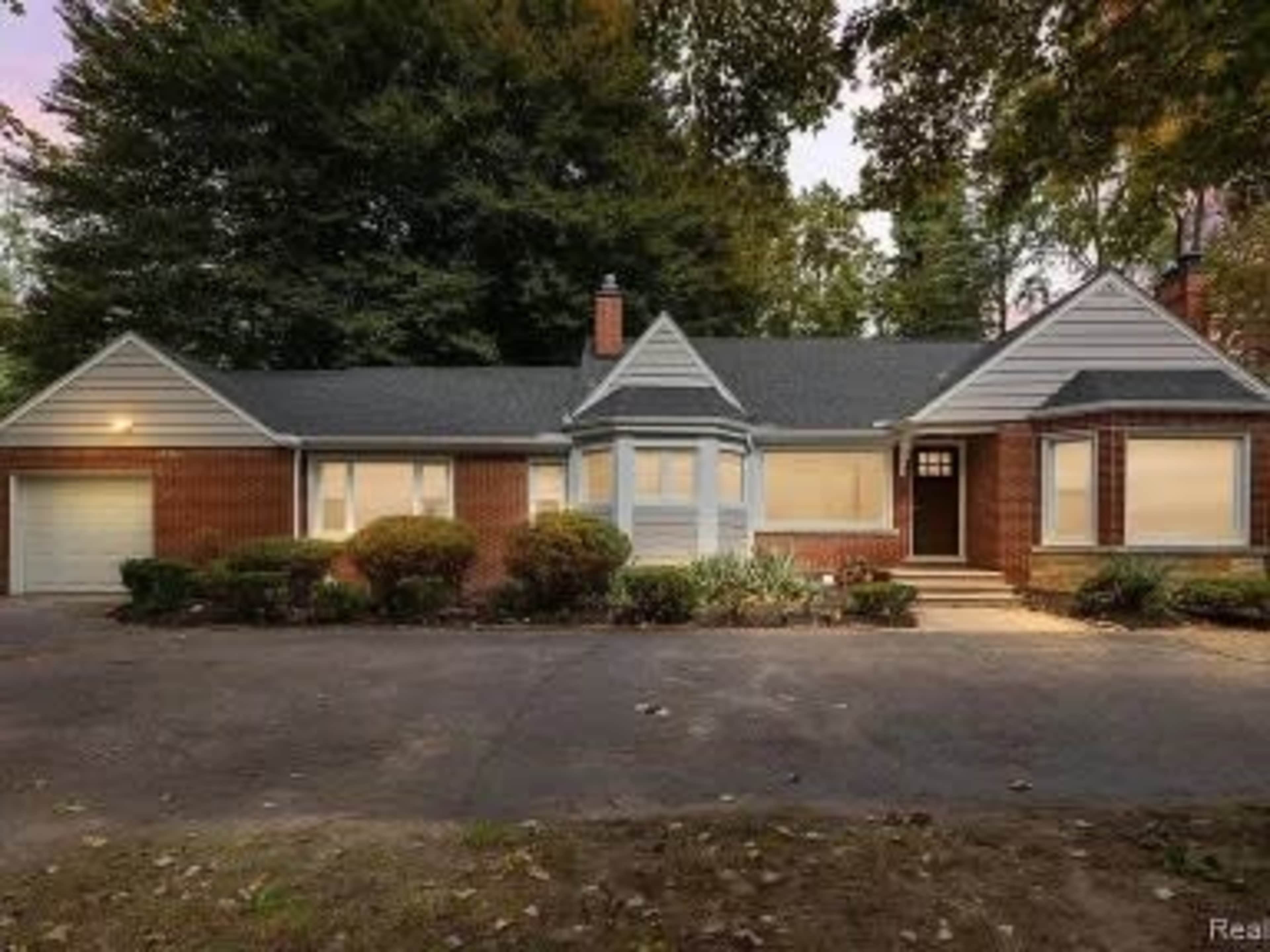 A single-story brick and siding house with a triangular roof and a front driveway surrounded by shrubbery.