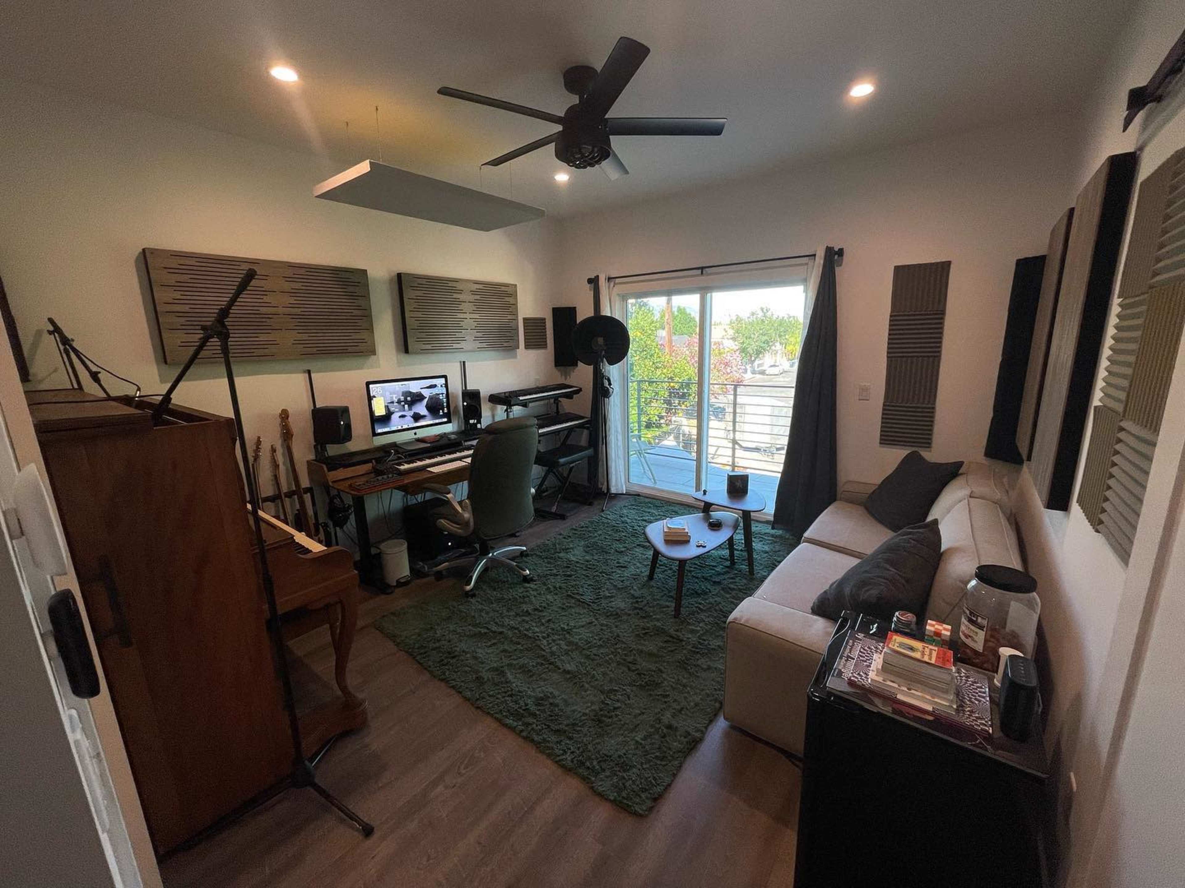 The image shows a music studio featuring a workspace with a computer setup, a couch, and soundproofing panels on the walls.