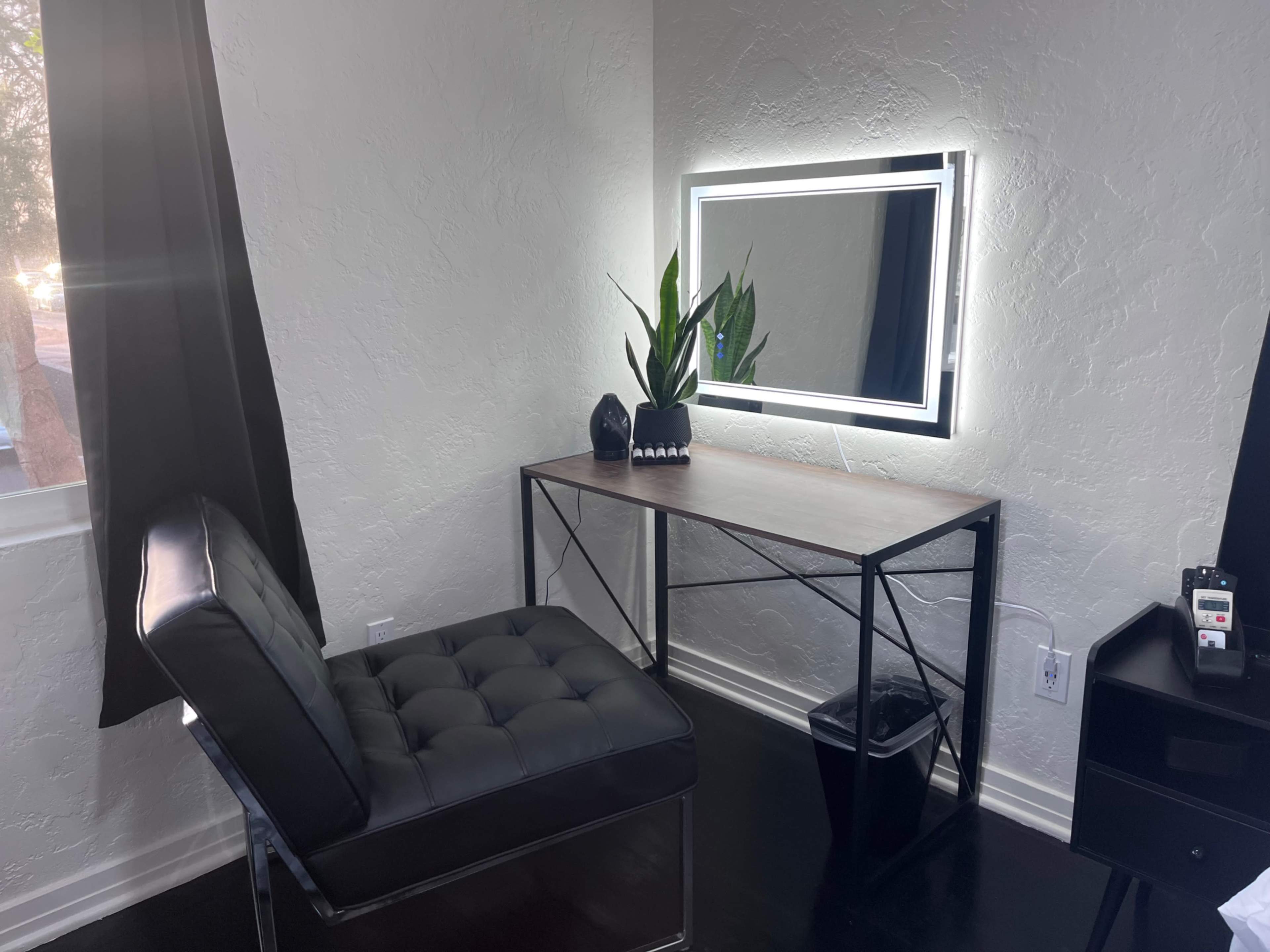 A modern vanity area features a sleek chair, a wooden desk with a lit mirror, and a potted plant against a light-colored wall.