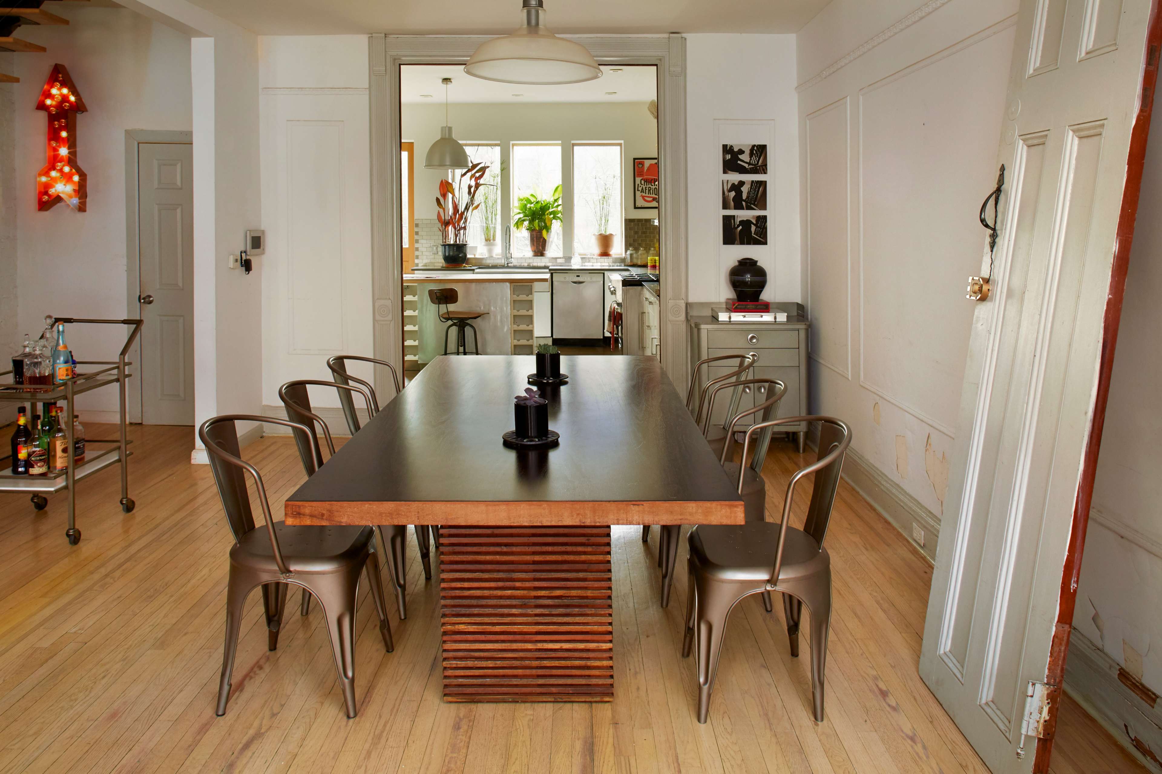 A long wooden dining table with metal chairs sits in a well-lit room, leading to a kitchen area with plants and decor visible in the background.
