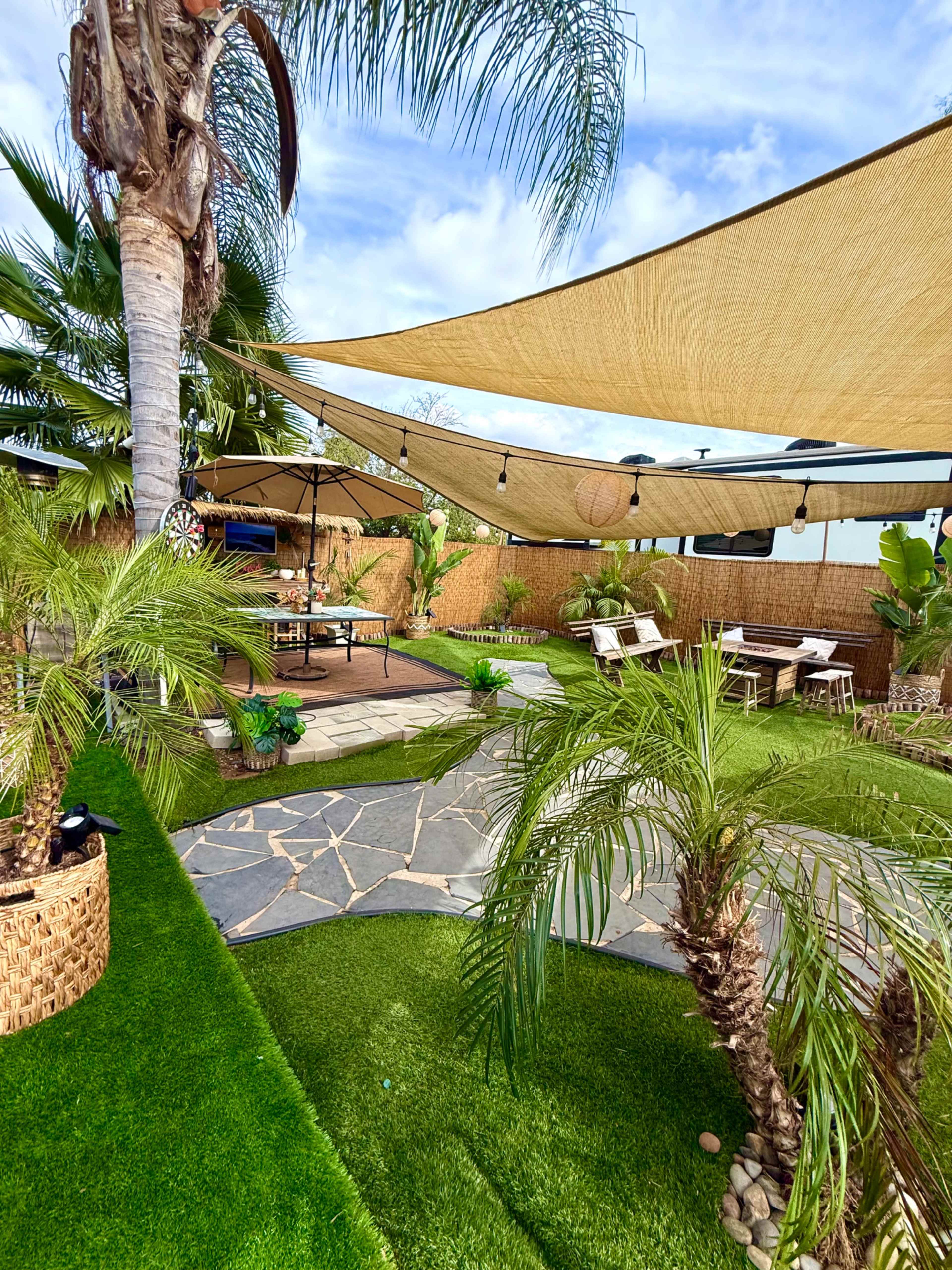 The image shows a landscaped backyard with green artificial grass, palm trees, and shaded seating areas under large fabric canopies.