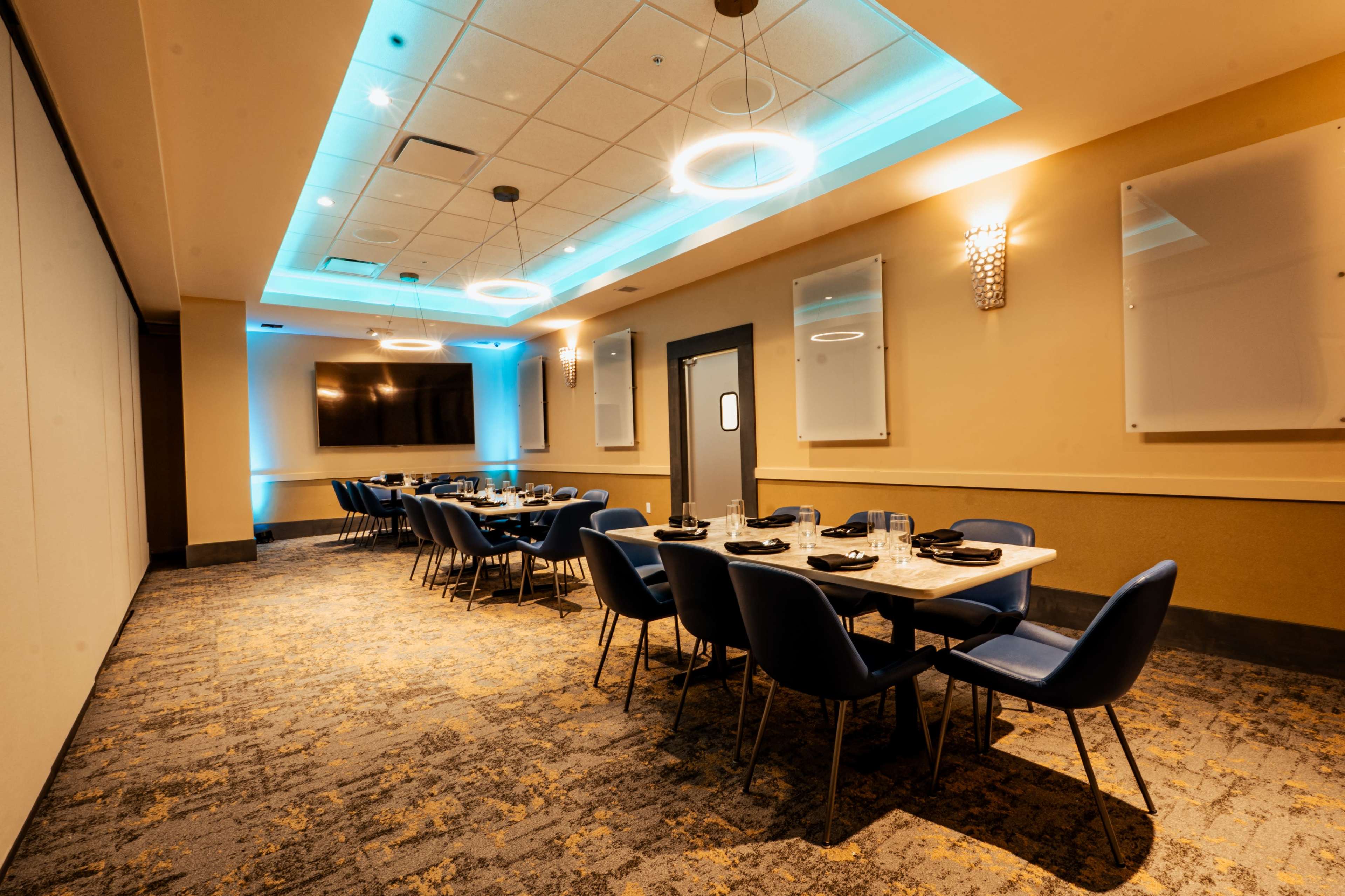 The image shows a conference room with a long table set for a meal, surrounded by blue chairs under modern lighting.