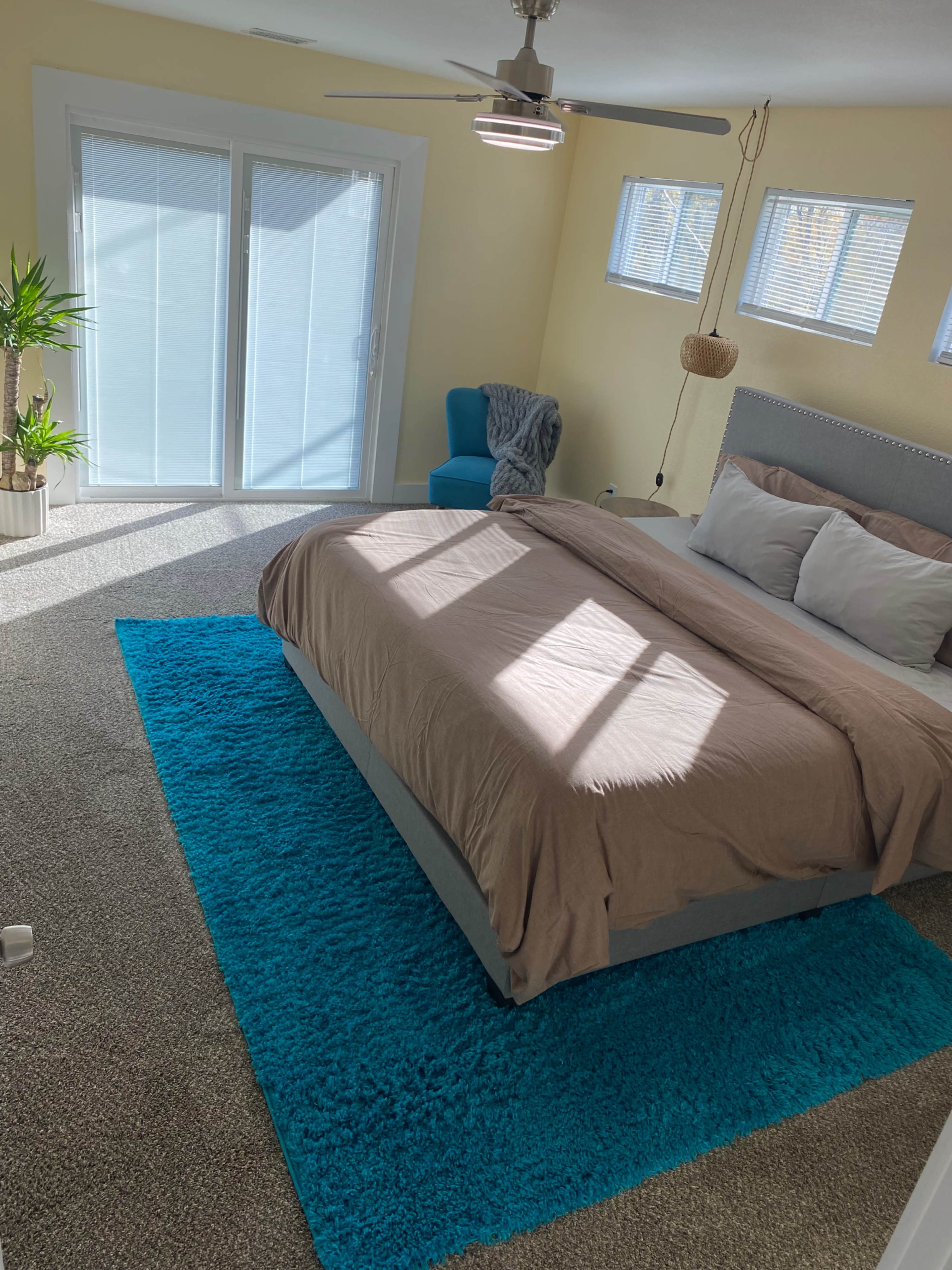 The image shows a well-lit bedroom with a bed covered in a tan blanket, a blue rug on carpet, and large sliding glass doors allowing natural light to enter.