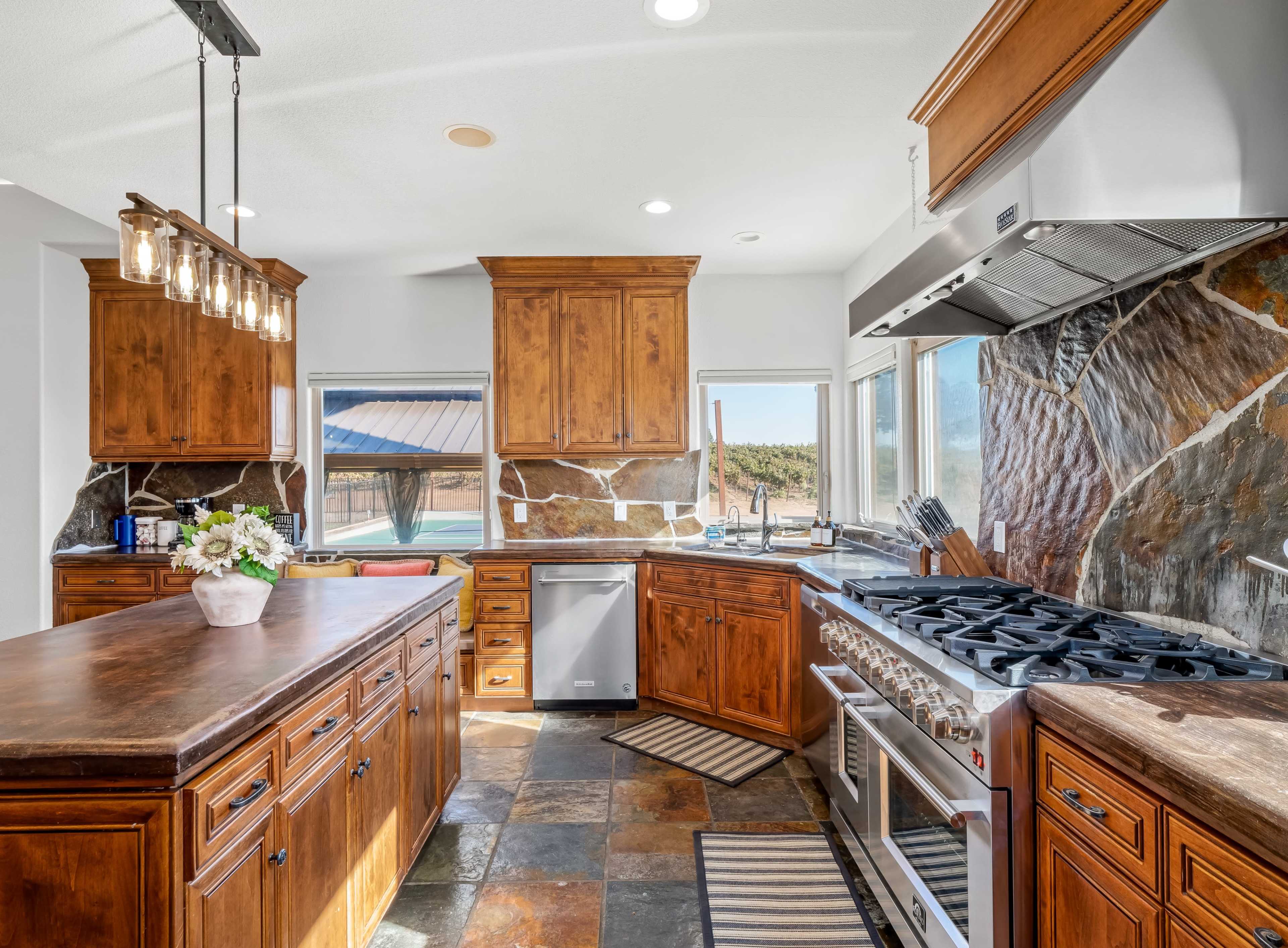 A kitchen features wooden cabinetry, a large island with a dark countertop, stainless steel appliances, and slate stone accents.
